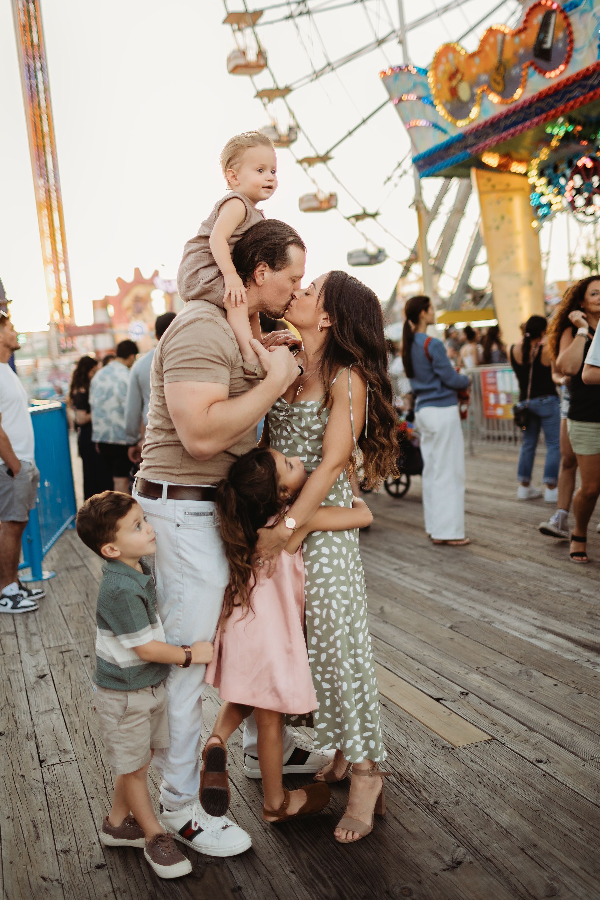 Family kissing on boardwalk, two children held close, one on father's shoulders; amusement park in background.