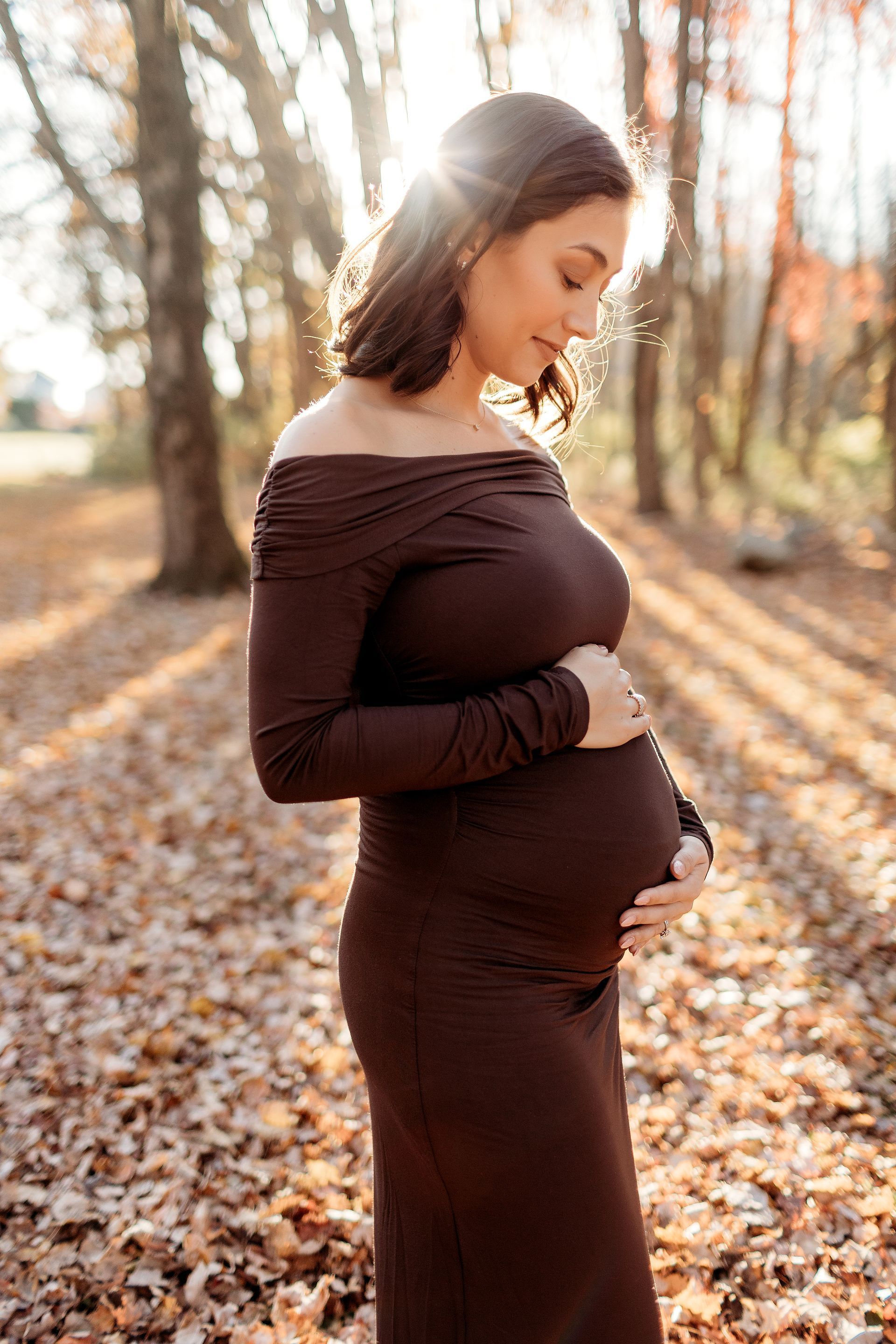 Pregnant woman in brown dress cradling belly, outdoors. Sunlight and fall foliage in background.
