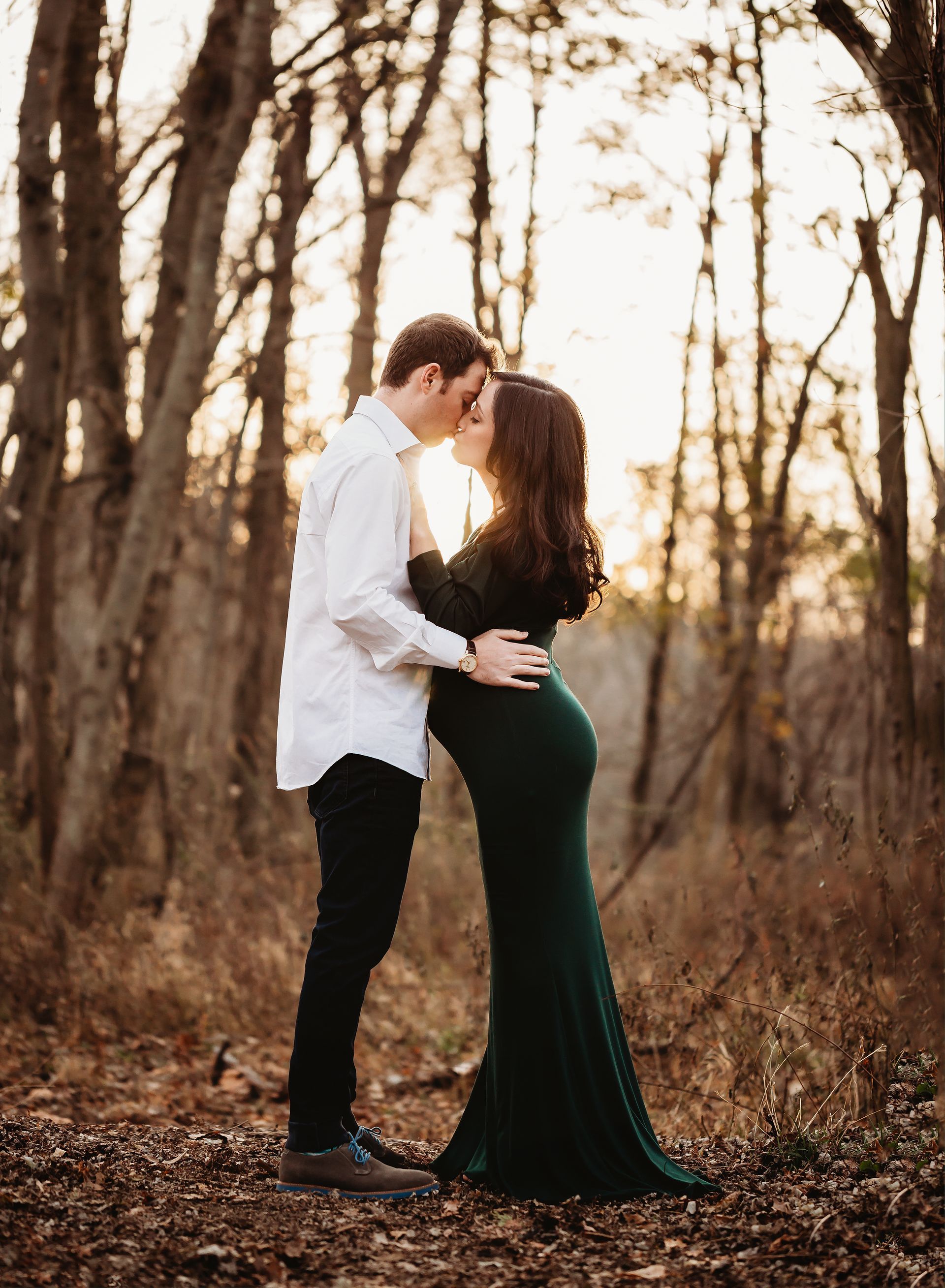 A man and a pregnant woman are kissing in the woods.
