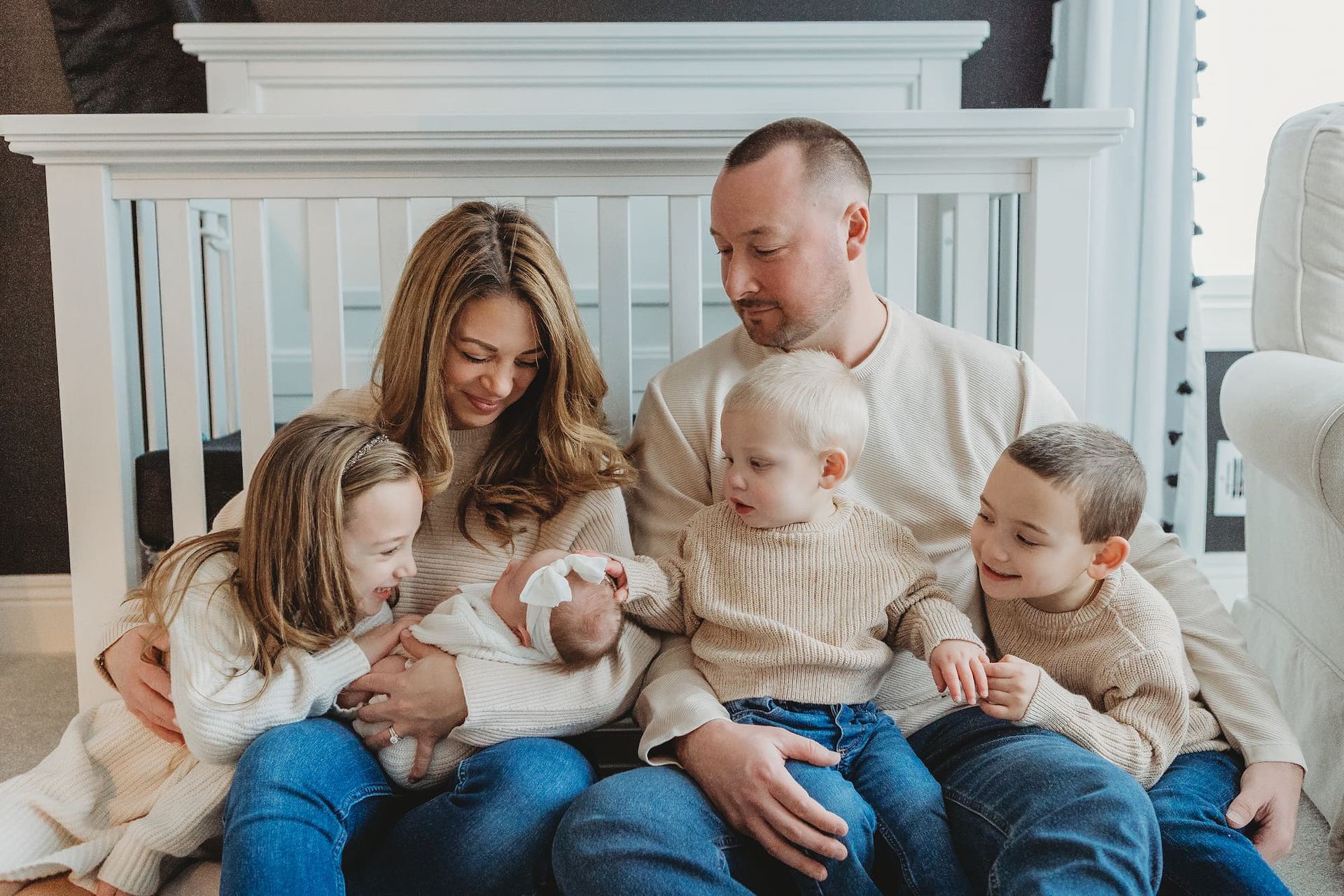 A family of six, dressed in neutral tones, sits together indoors, holding their newborn baby while smiling softly in Glen Rock, NJ.