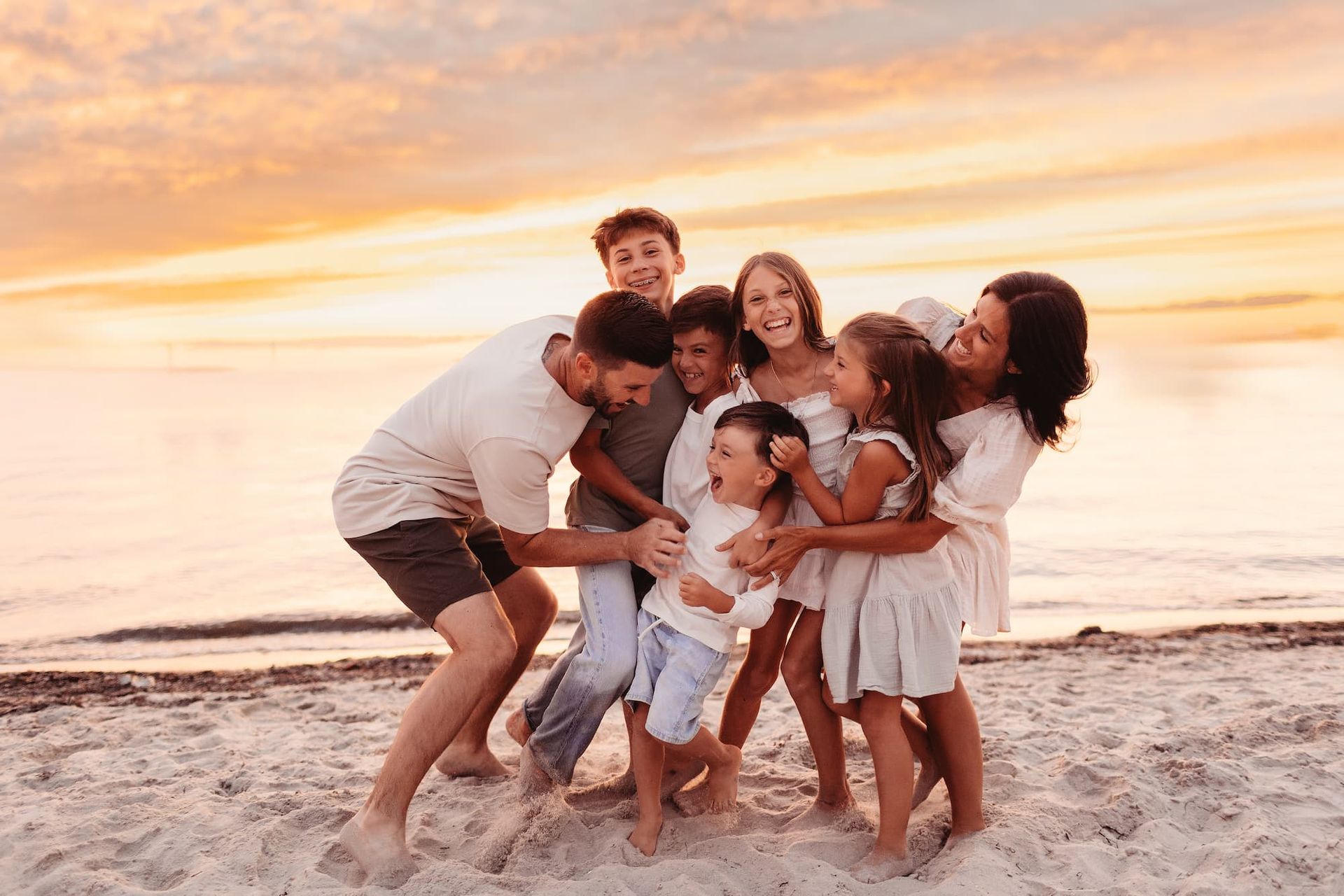 Family hugs on beach at sunset. People smile, laugh, and embrace on the sand with the ocean in the background.