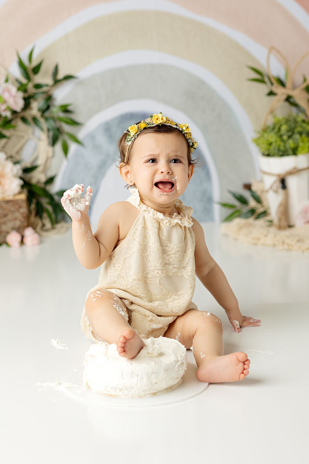 Baby in a cream romper and floral crown sits in front of a rainbow backdrop, covered in flour, crying.