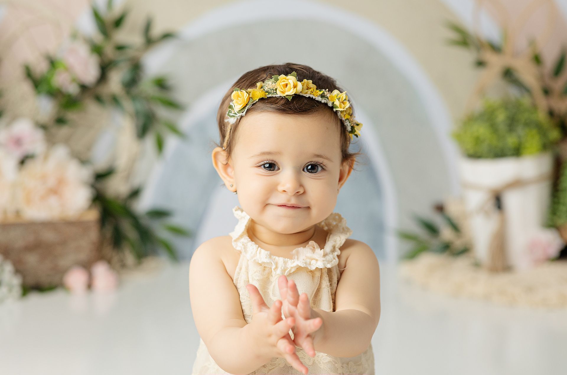 Smiling baby wearing a yellow floral headband, clapping hands, in front of a neutral-toned backdrop with flowers.