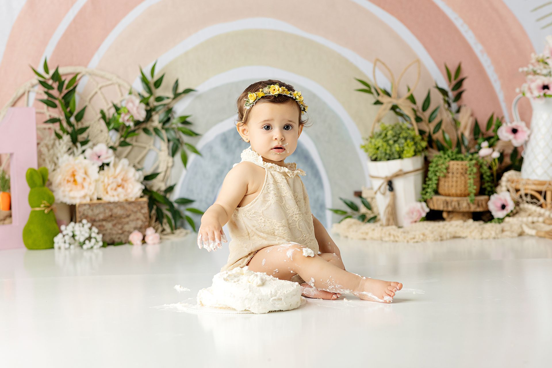 Baby sits amidst cake, wearing floral headband, in front of a rainbow backdrop with flowers.