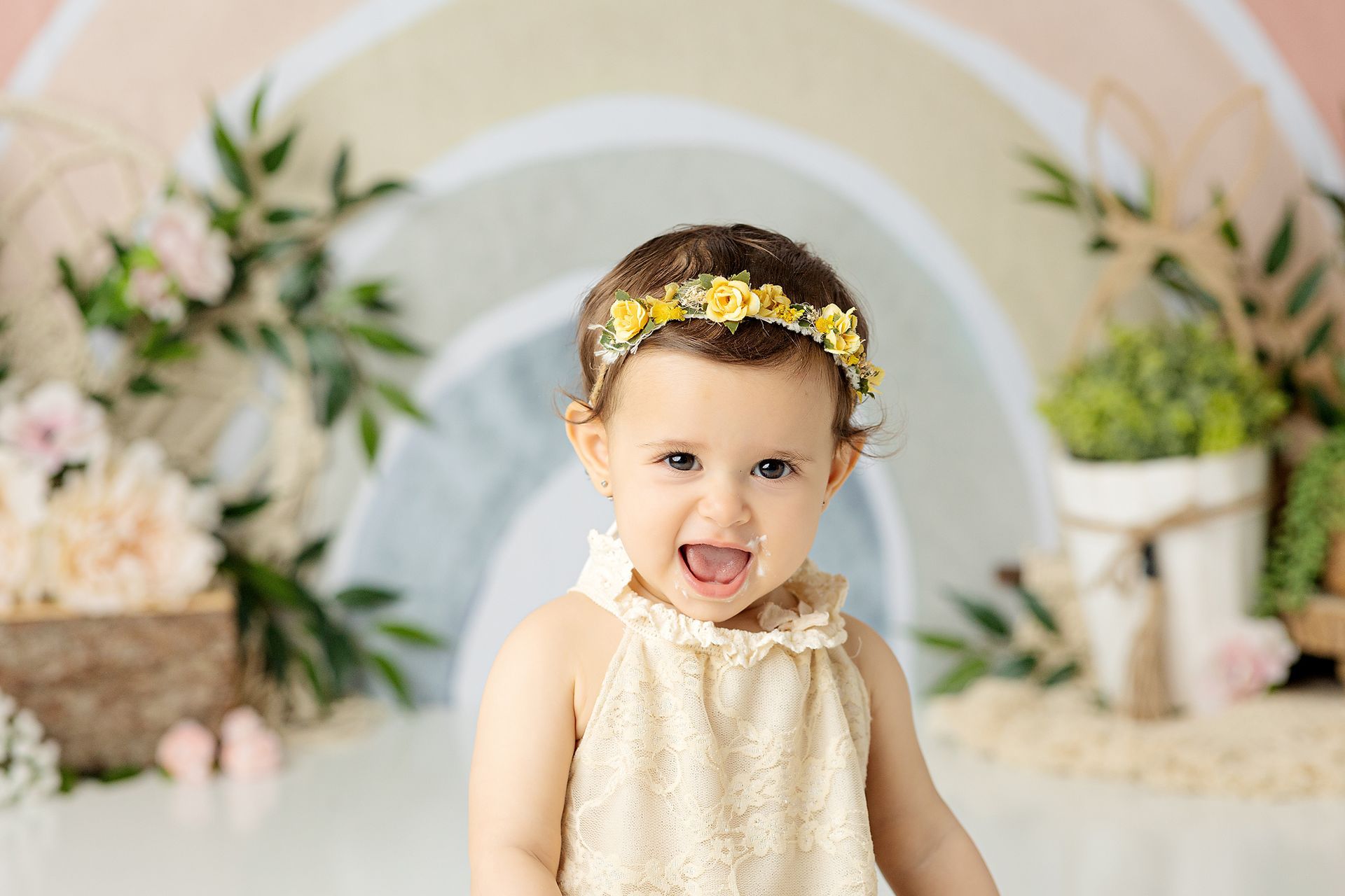 Toddler with flower crown, smiling, open mouth; pastel rainbow backdrop, floral accents.