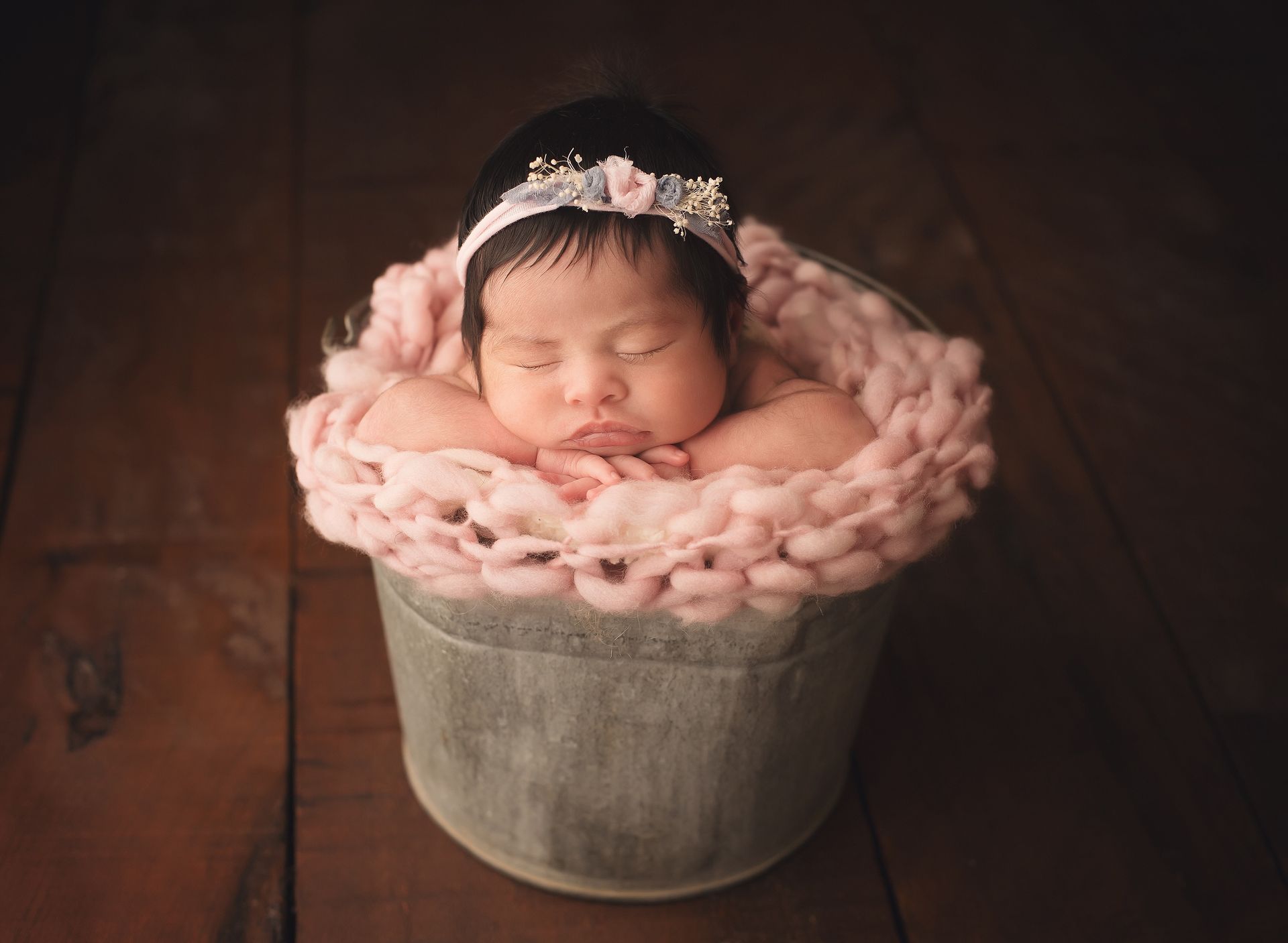 A newborn baby is sleeping in a bucket on a wooden floor.