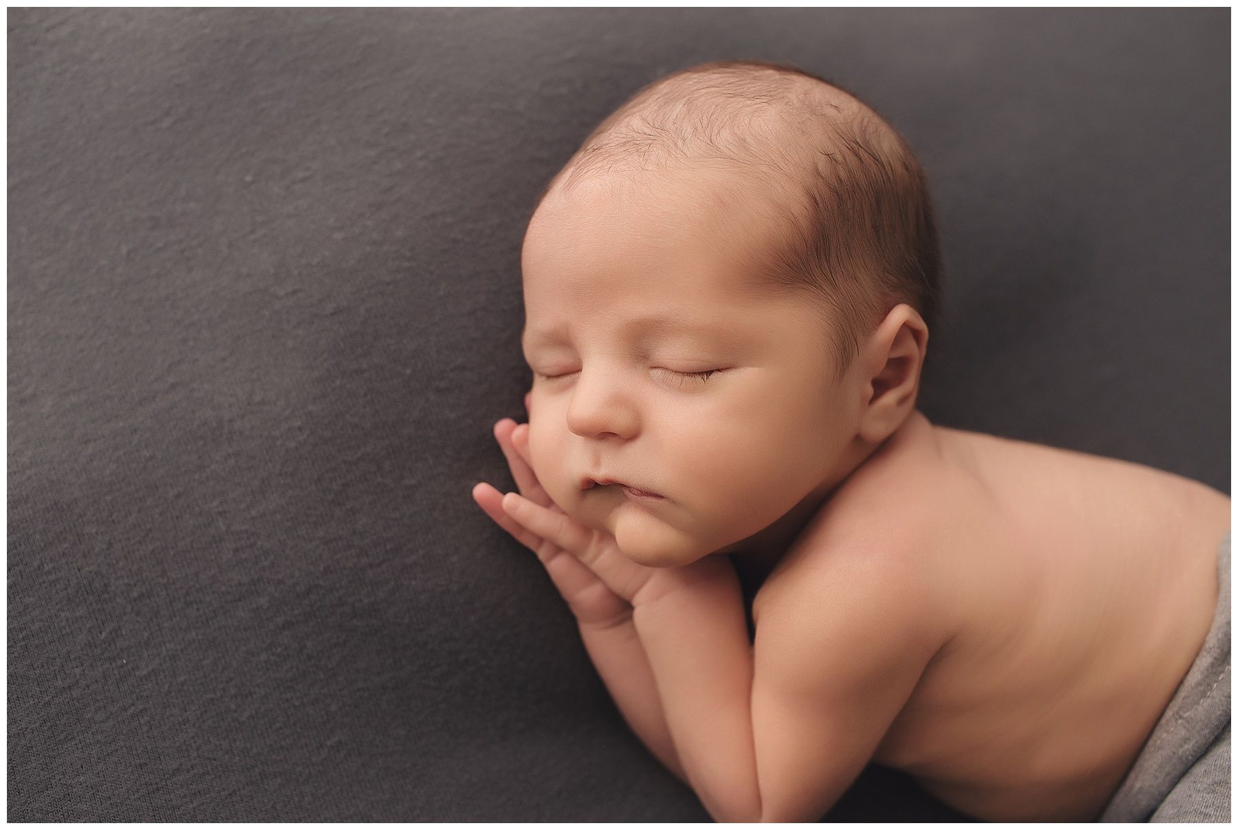 A newborn baby is sleeping on a gray blanket with his hands on his face.