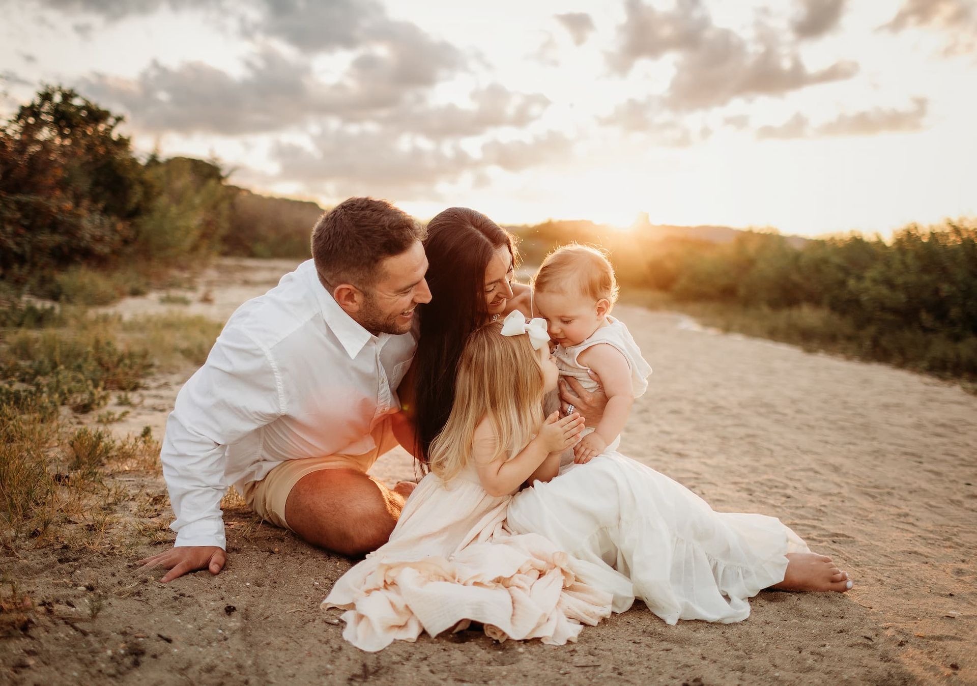 Family of four sits on sand, mother holds two children, father looks on; sunset background.