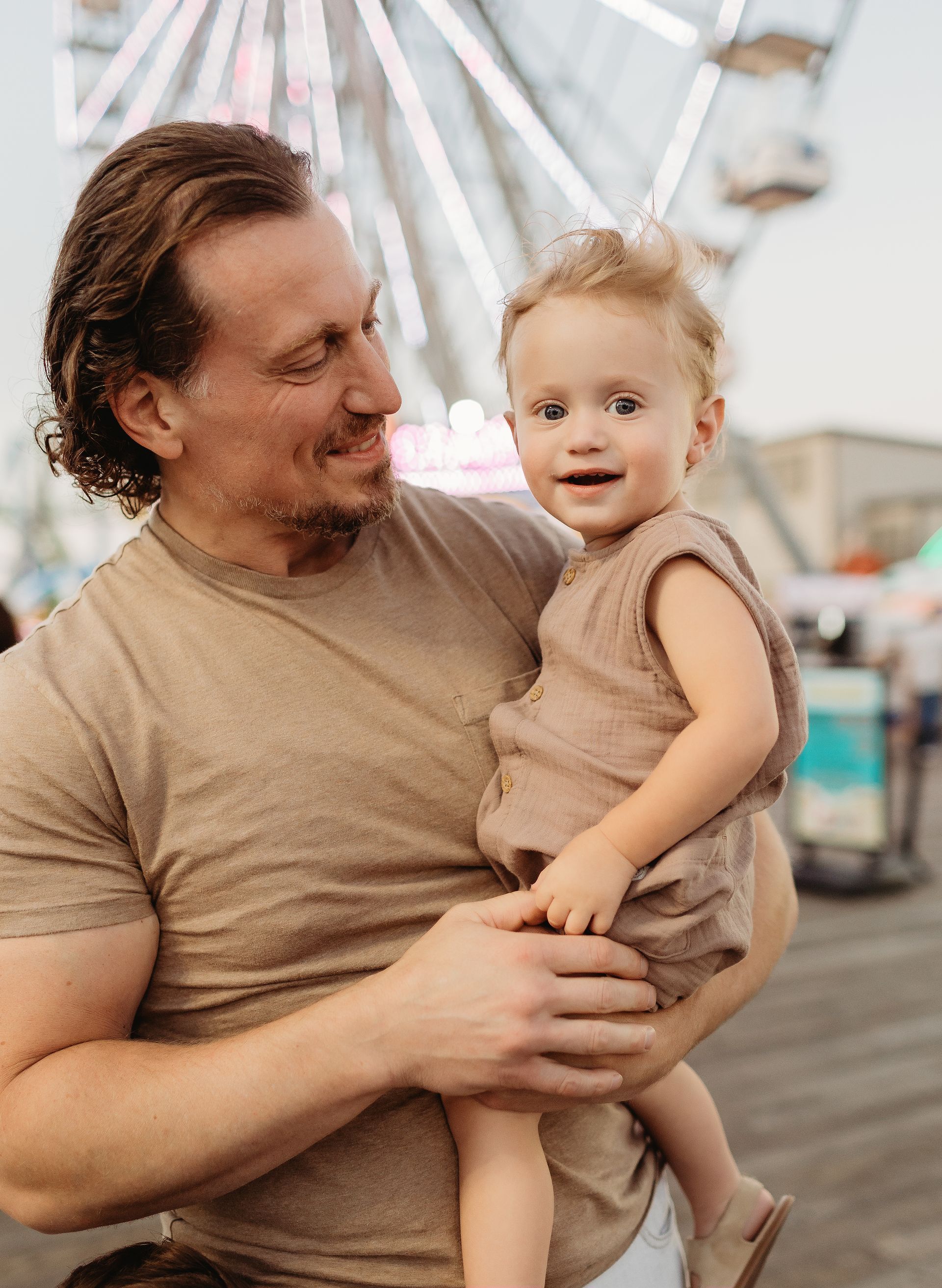 Man holding a smiling baby at an amusement park, Ferris wheel in the background.