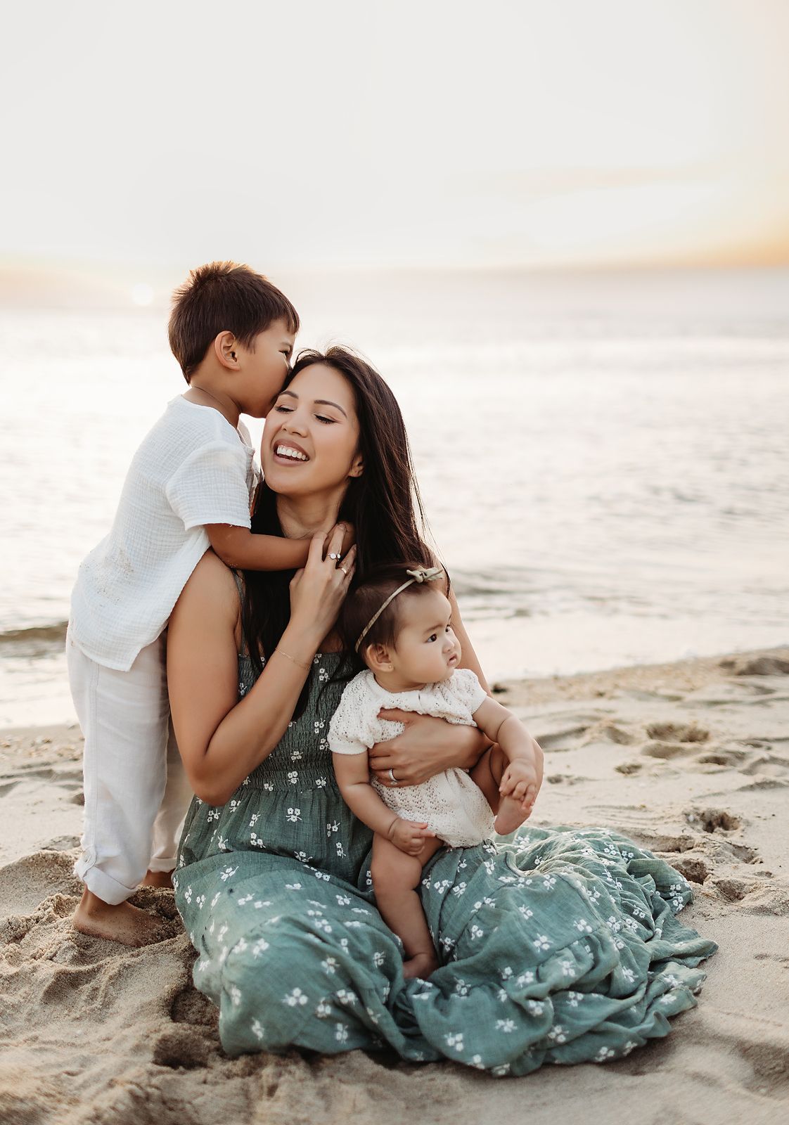 Woman on beach with two children; son kissing her, baby in her arms.