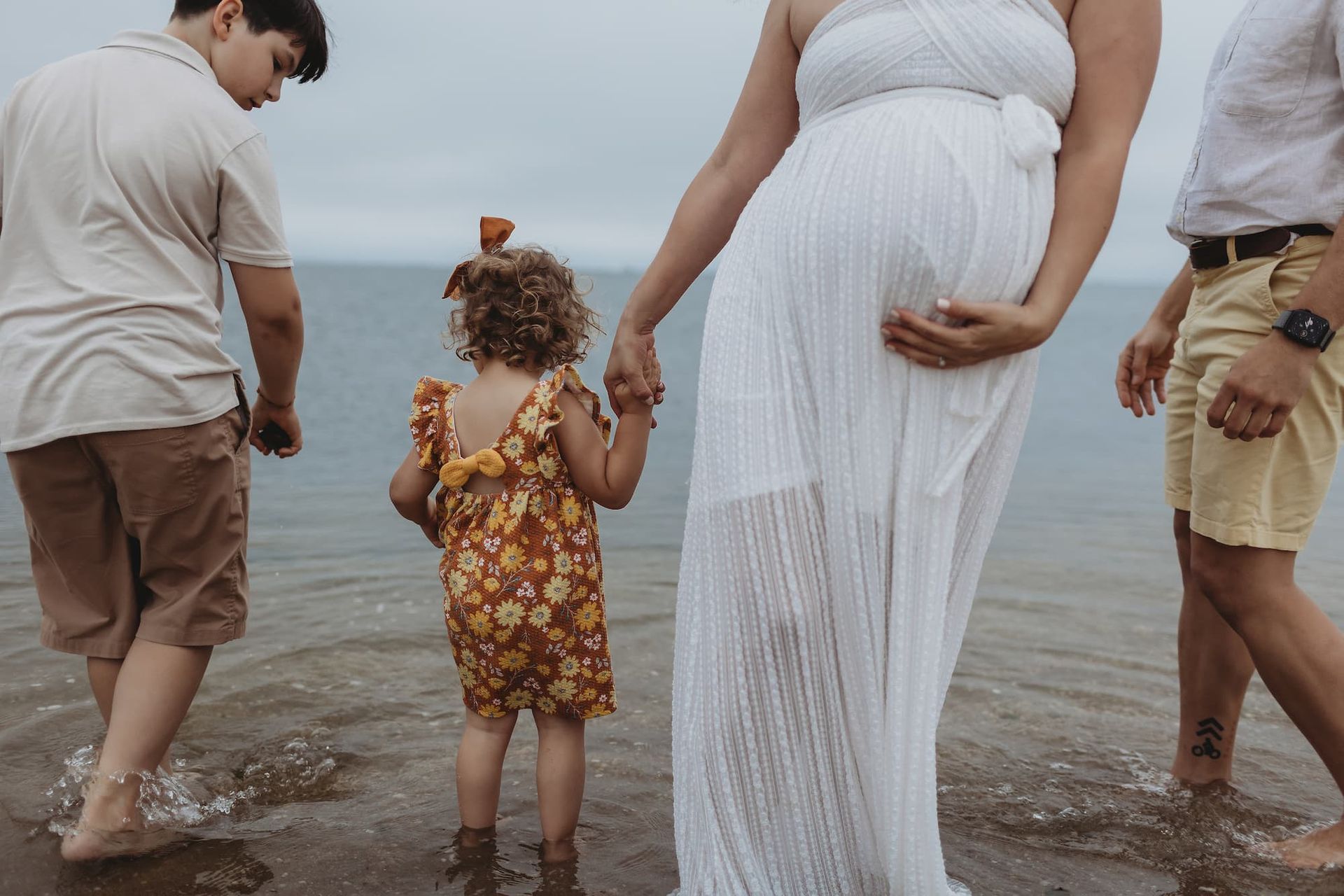 Family wading in shallow water: pregnant person in white dress holds hands with child; two others nearby.