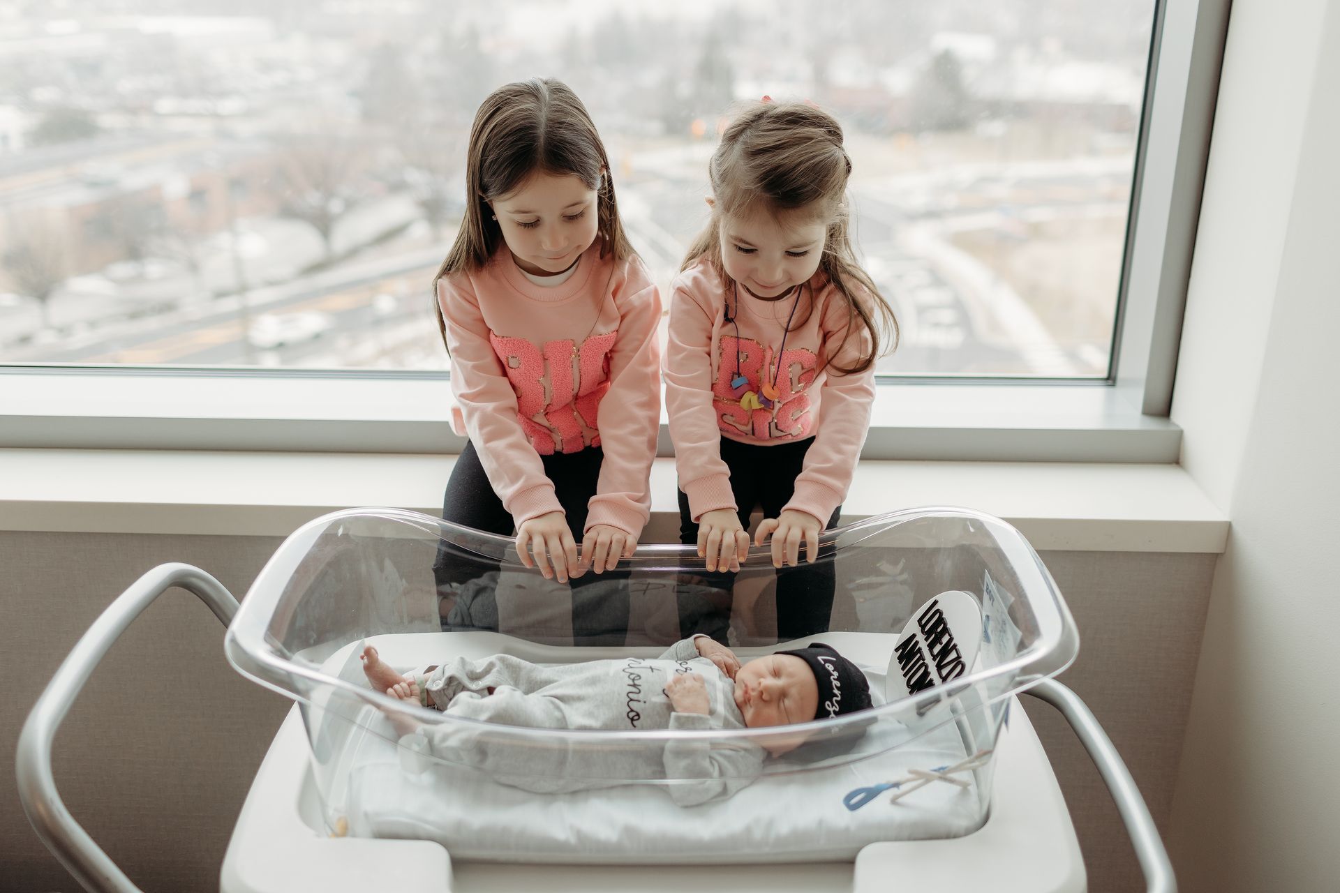 Two little girls are looking at a newborn baby in a hospital crib.