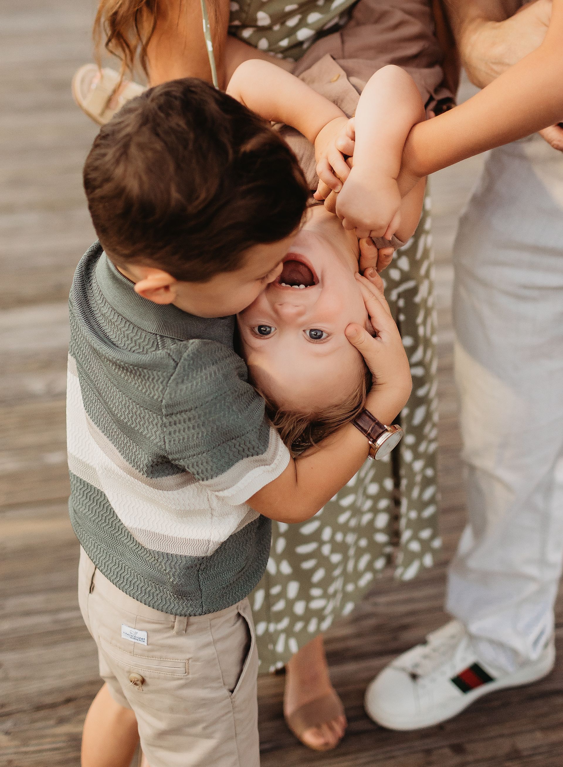 Boy kisses a baby held upside down by family; joyful expressions.