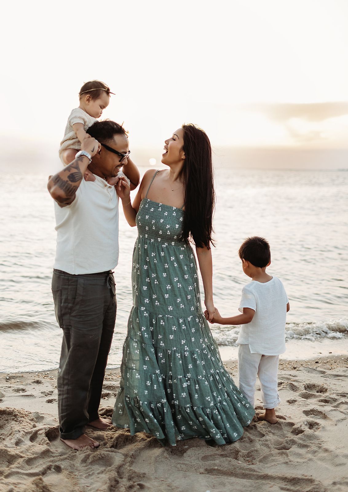 Family on beach at sunset: parents laughing, holding hands with children; one child on dad's shoulders.