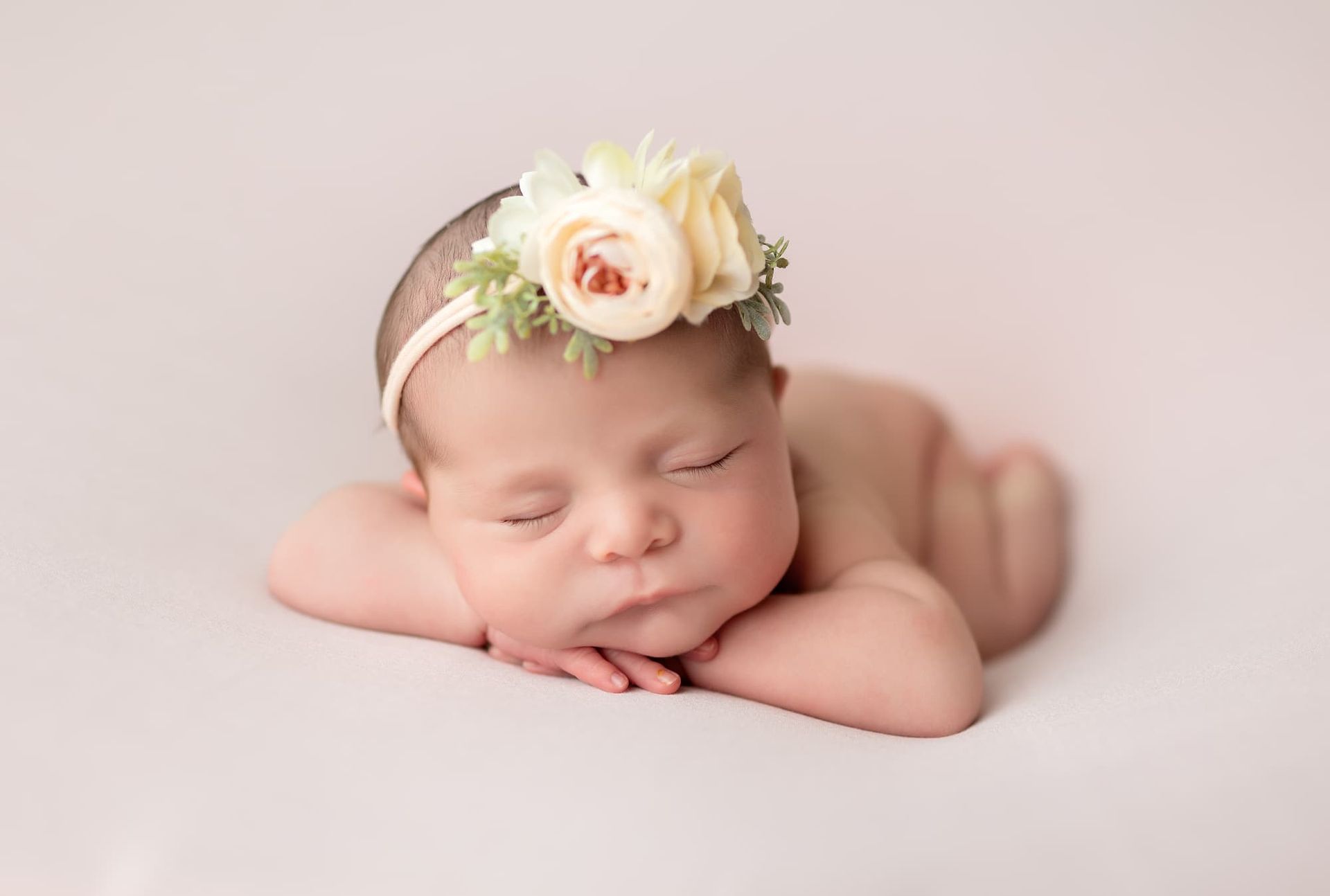 Newborn baby sleeping, wearing a floral headband, resting on arms, pink background.