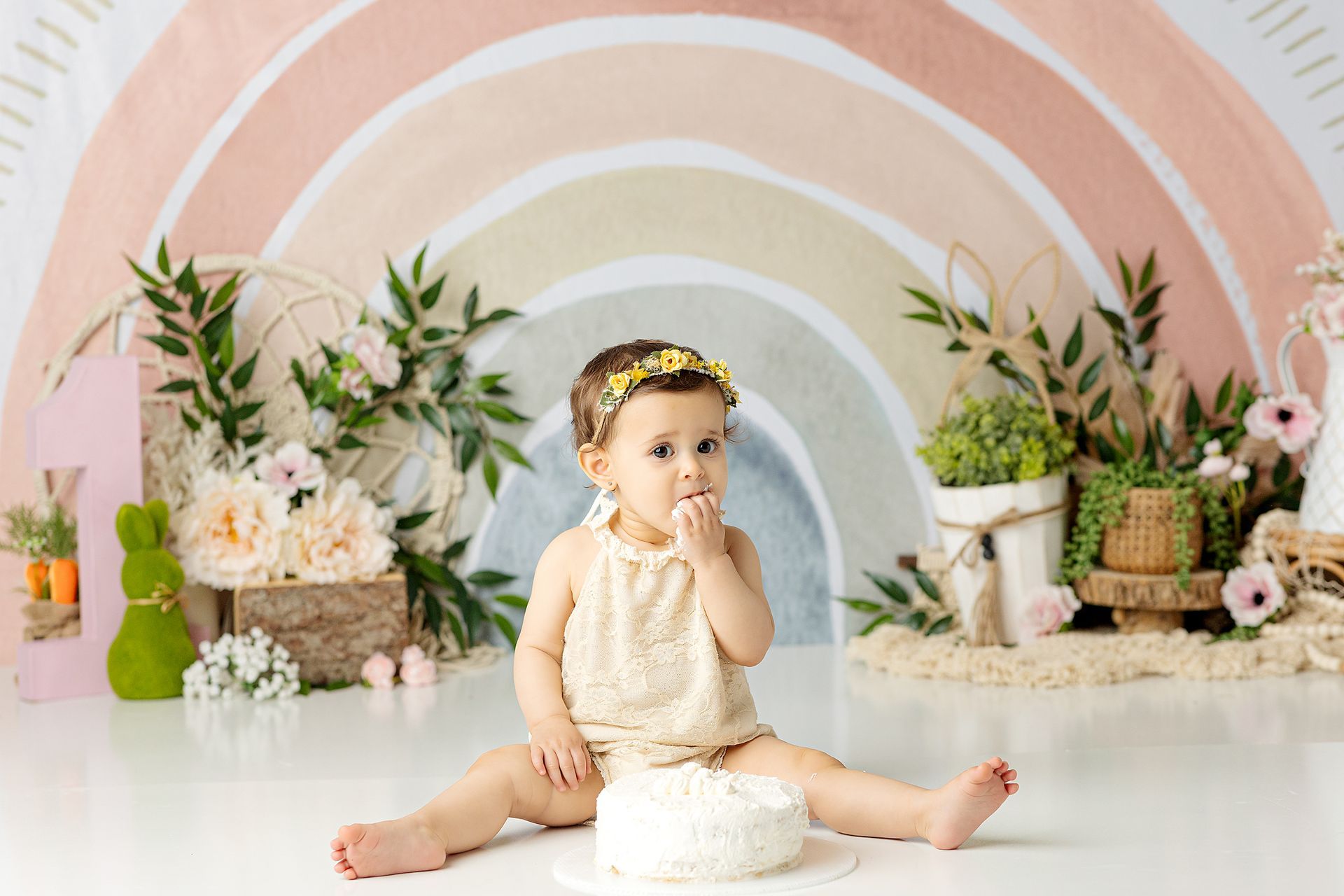Baby sits with cake, hand raised, floral crown, rainbow backdrop, surrounded by greenery.