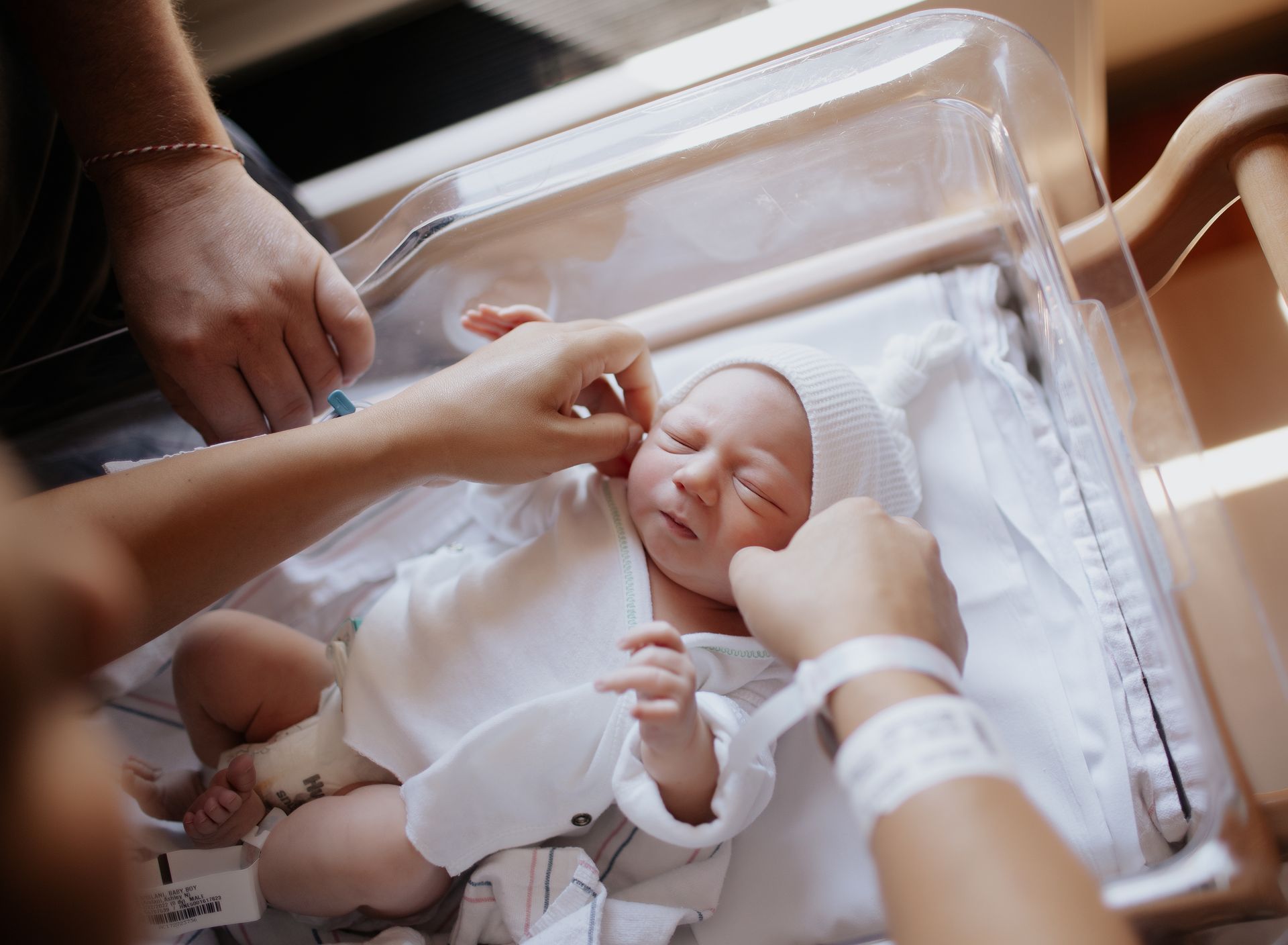 Newborn in a hospital bassinet, being examined by hands, wearing a white hat and outfit.