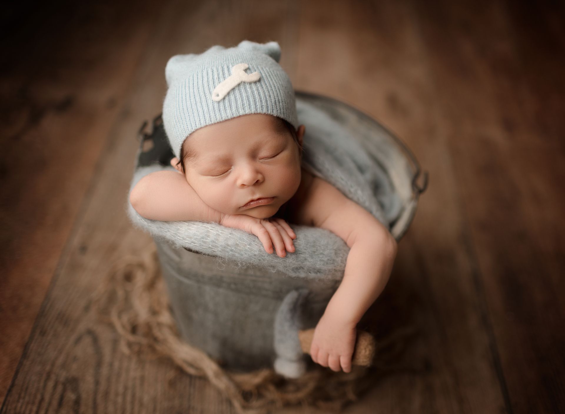 Newborn baby sleeping in a bucket, wearing a blue hat with a wrench.