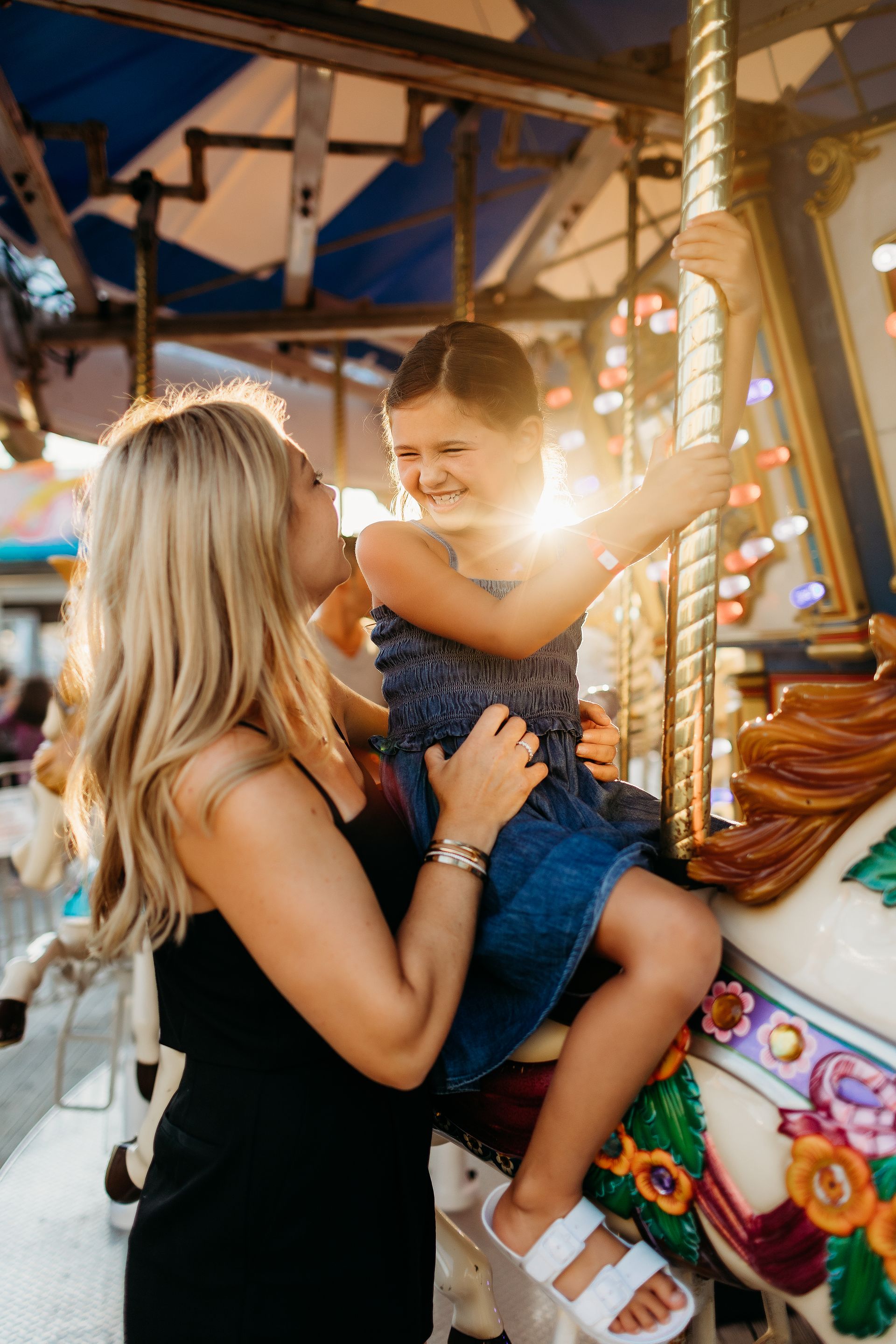 Woman helps child ride a carousel; setting sun in background.