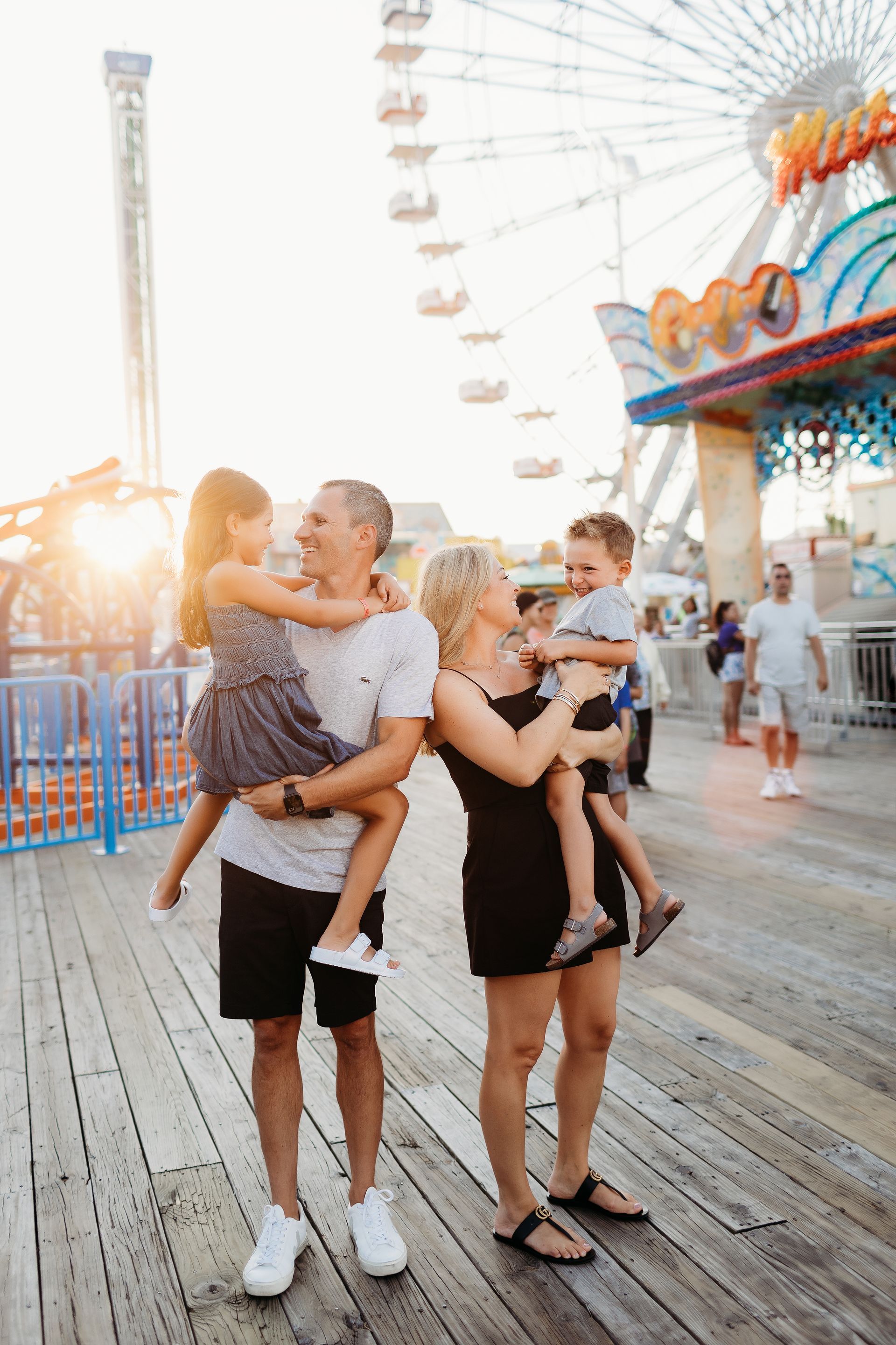Family of four on a boardwalk, holding children. Ferris wheel and setting sun in the background.