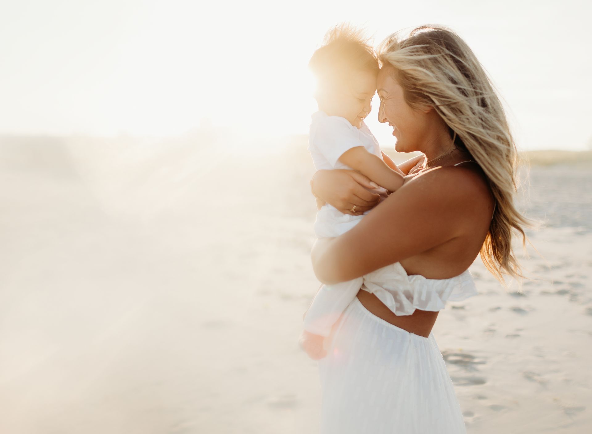 Woman holding a child, faces touching, on a beach; sunlit and warm.