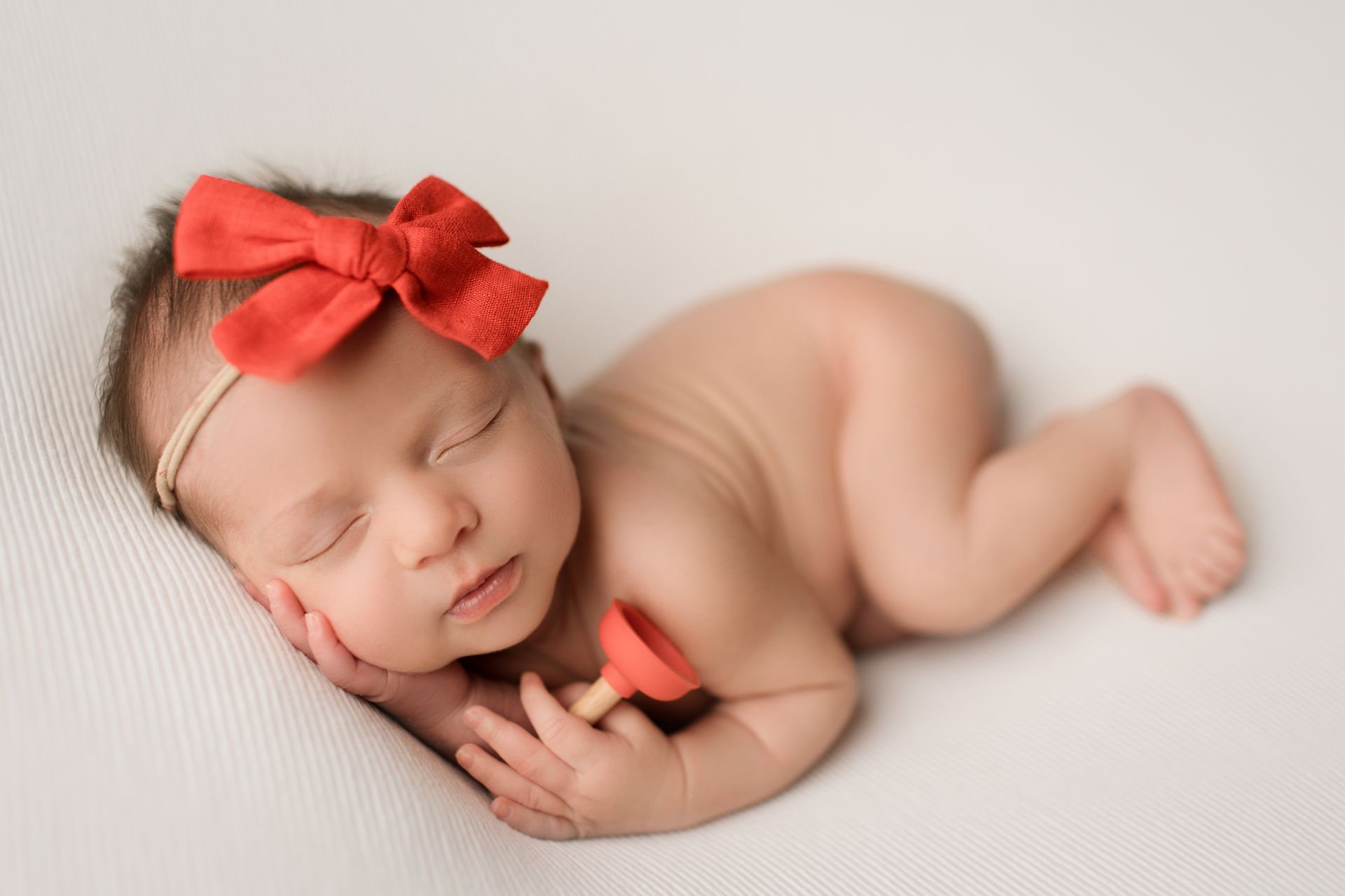 Newborn sleeping on a white blanket, wearing a red bow; holding a small plunger.