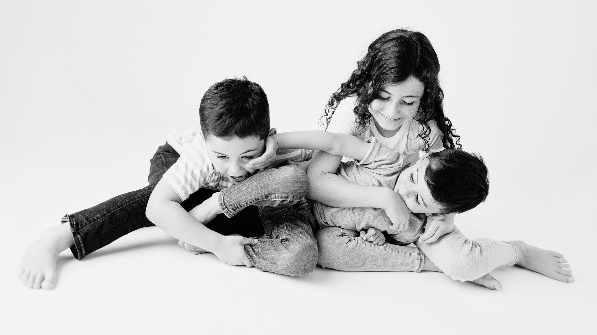 Three children playfully interacting against a white background; one leans on a fluffy bear, the others reach for each other.
