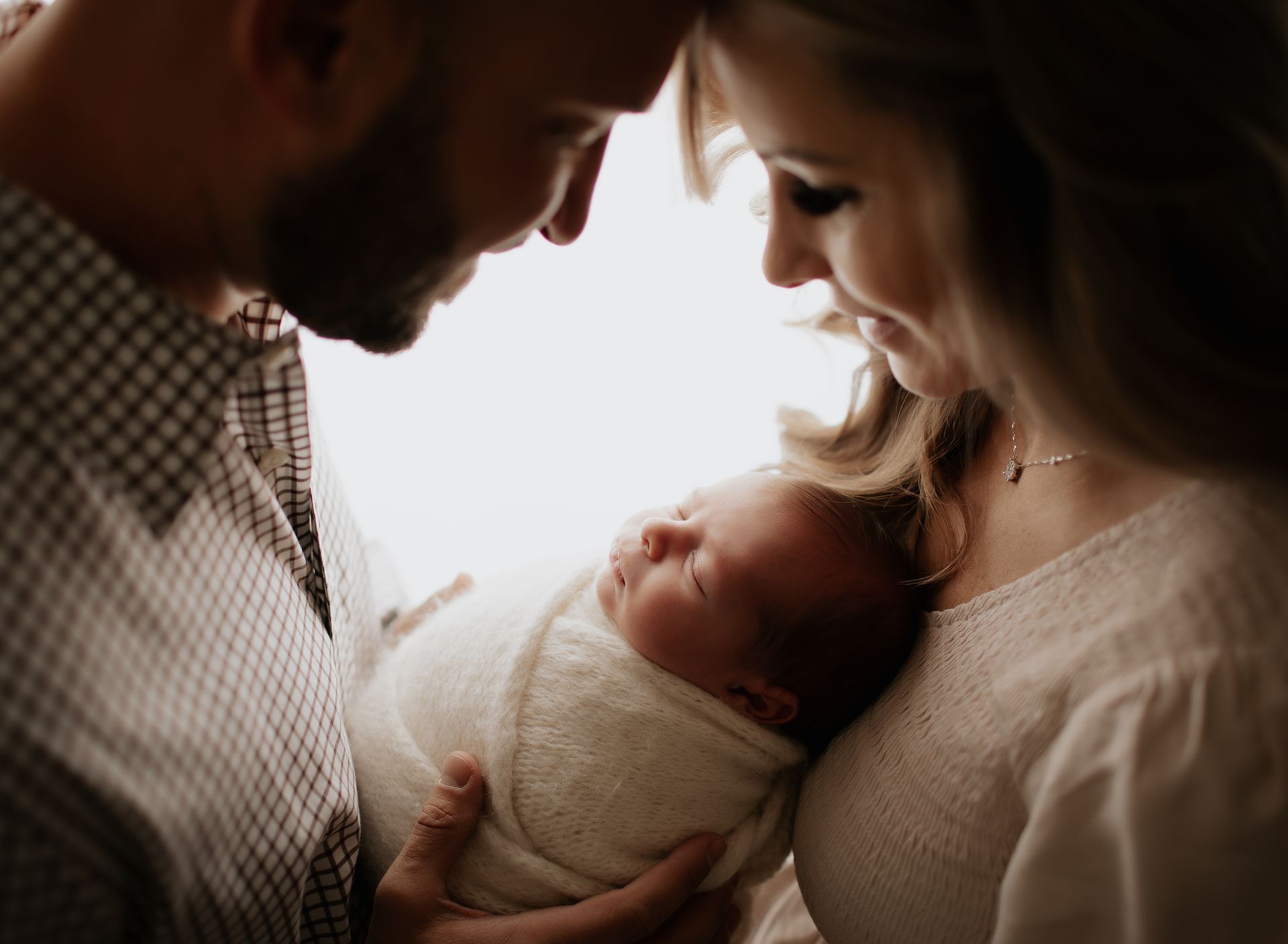 Parents gazing at a swaddled newborn baby, indoors. Soft lighting, neutral tones.