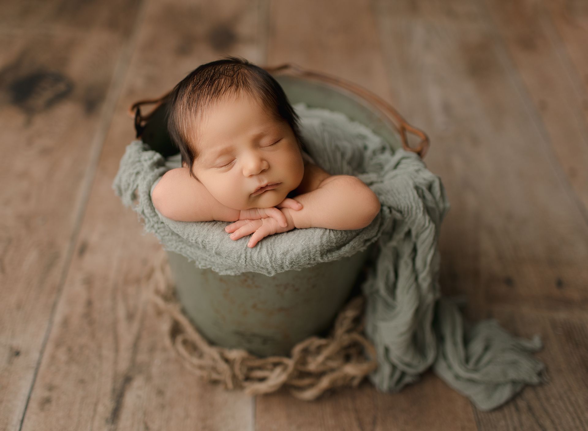 Newborn baby sleeping peacefully in a bucket lined with green fabric, resting on a wooden floor.