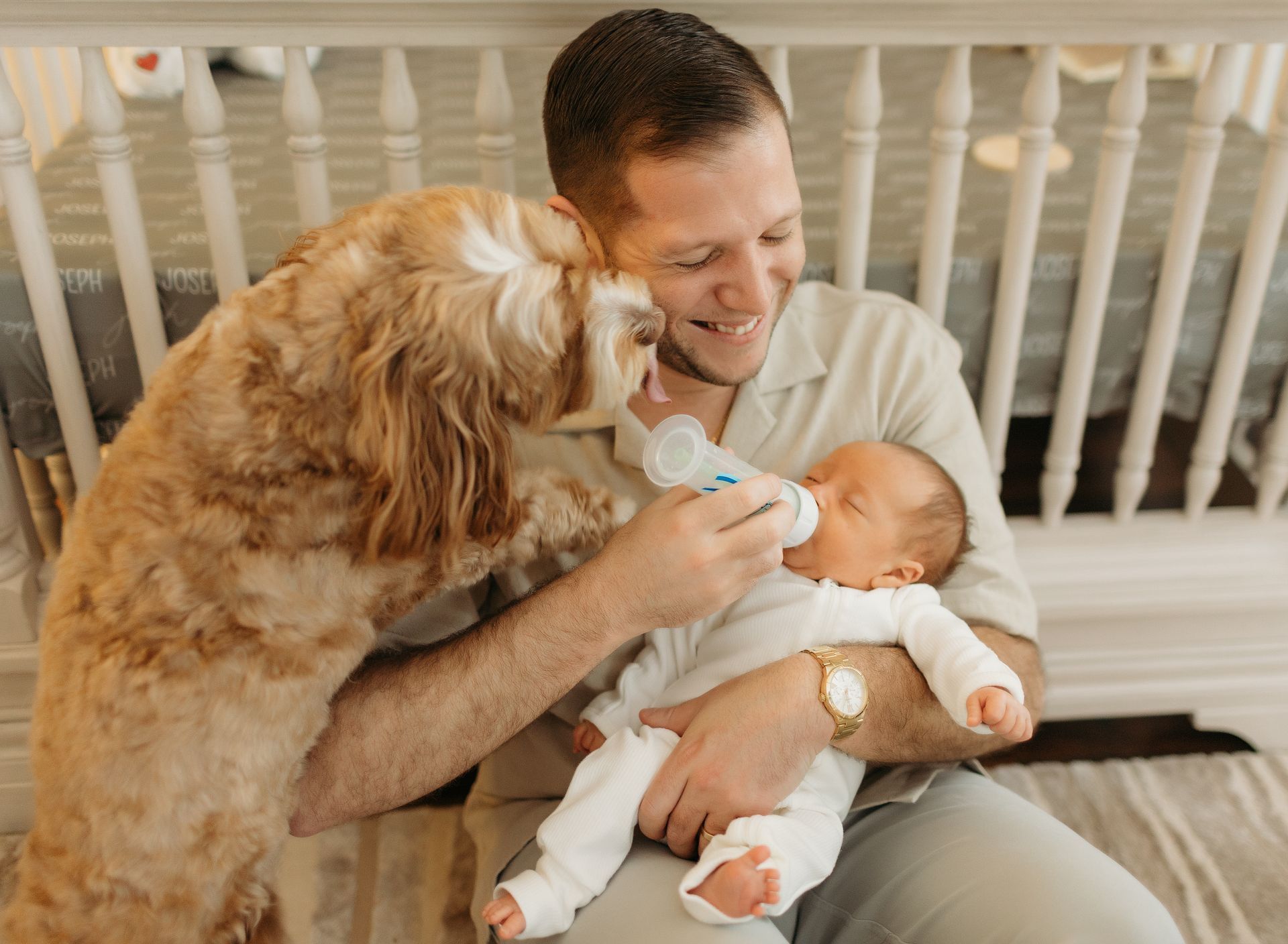 A man is feeding a baby from a bottle while a dog looks on.