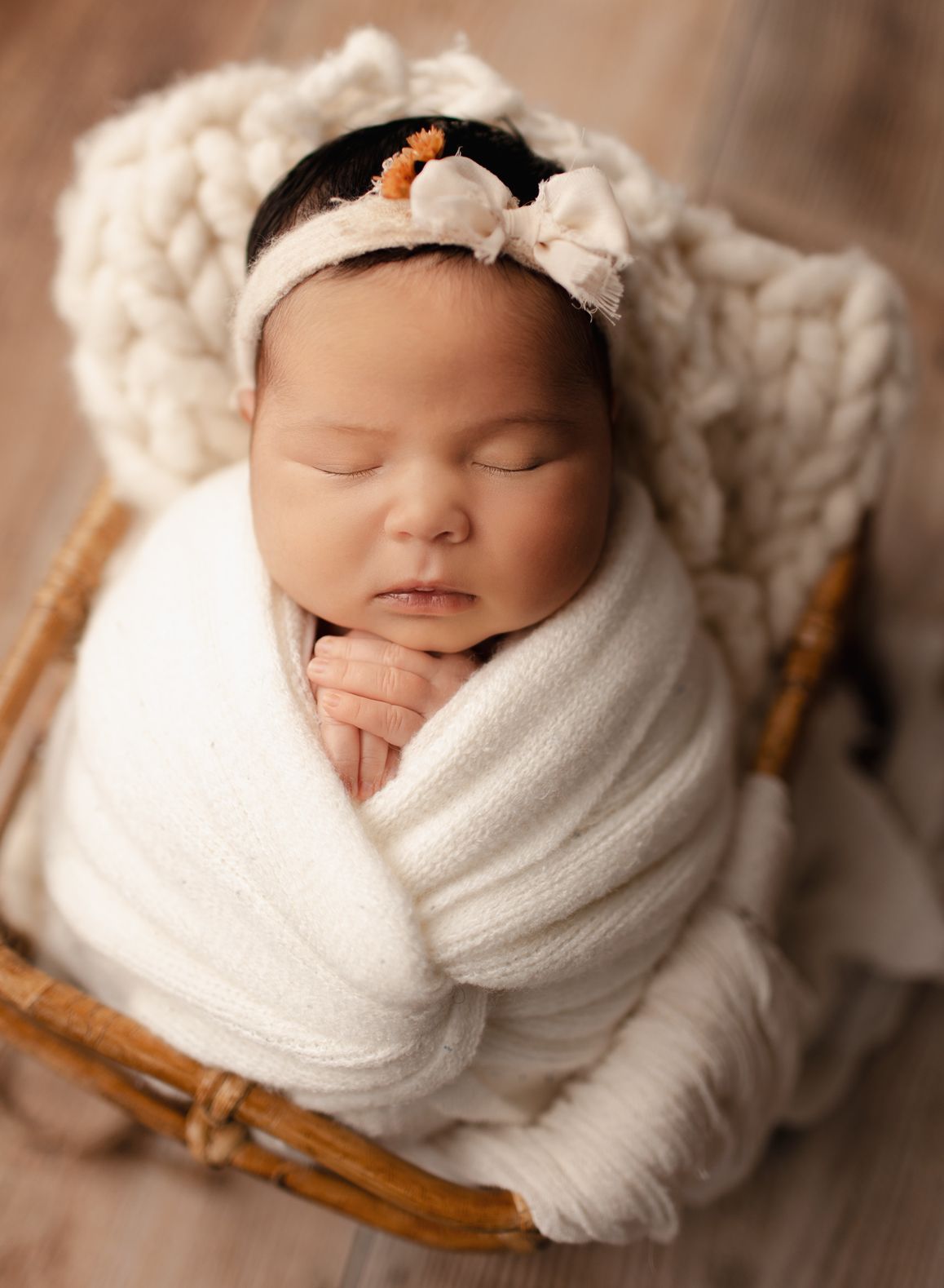 Newborn baby wrapped in white blanket, wearing a bow headband, in a woven basket.