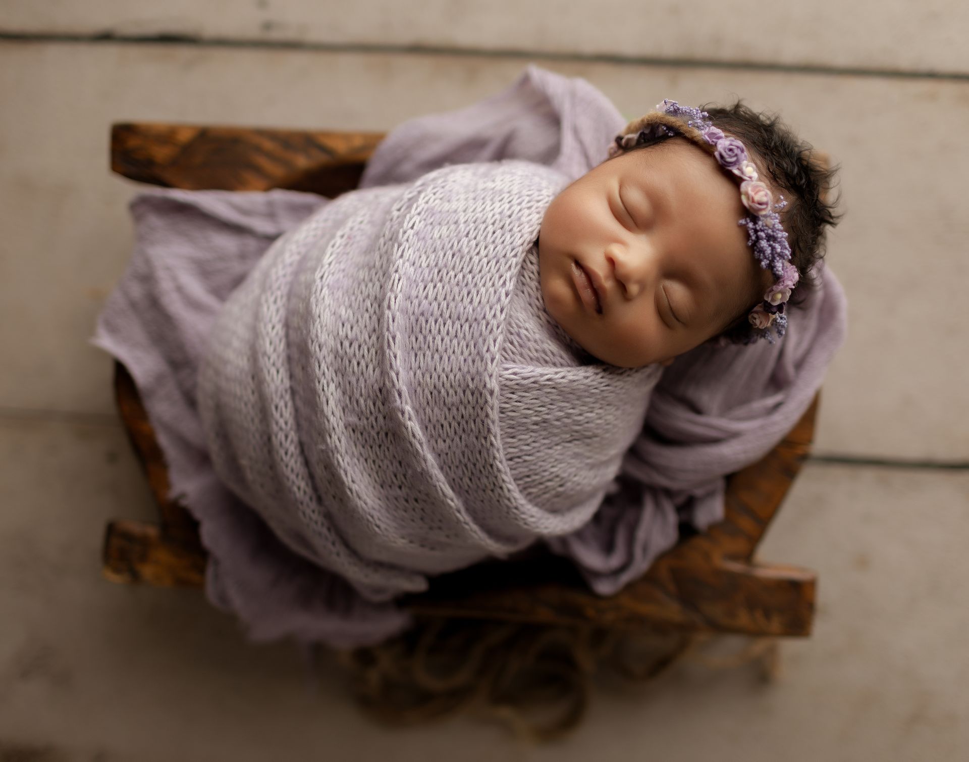 Newborn swaddled in purple blanket, wearing floral headband, in wooden cradle.