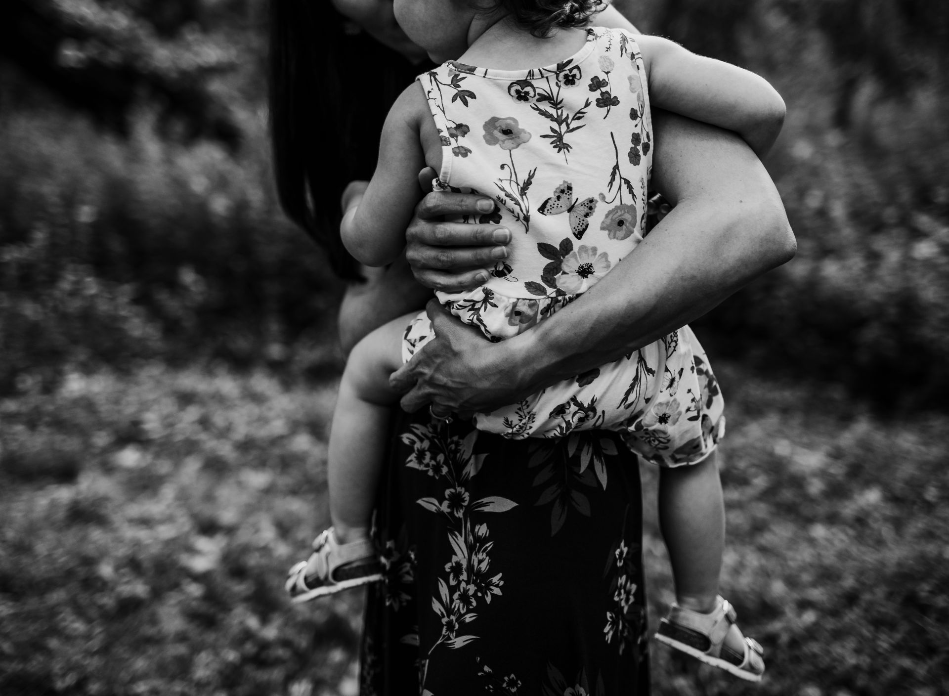 Person holding a toddler in a floral romper outside. Black and white photo.