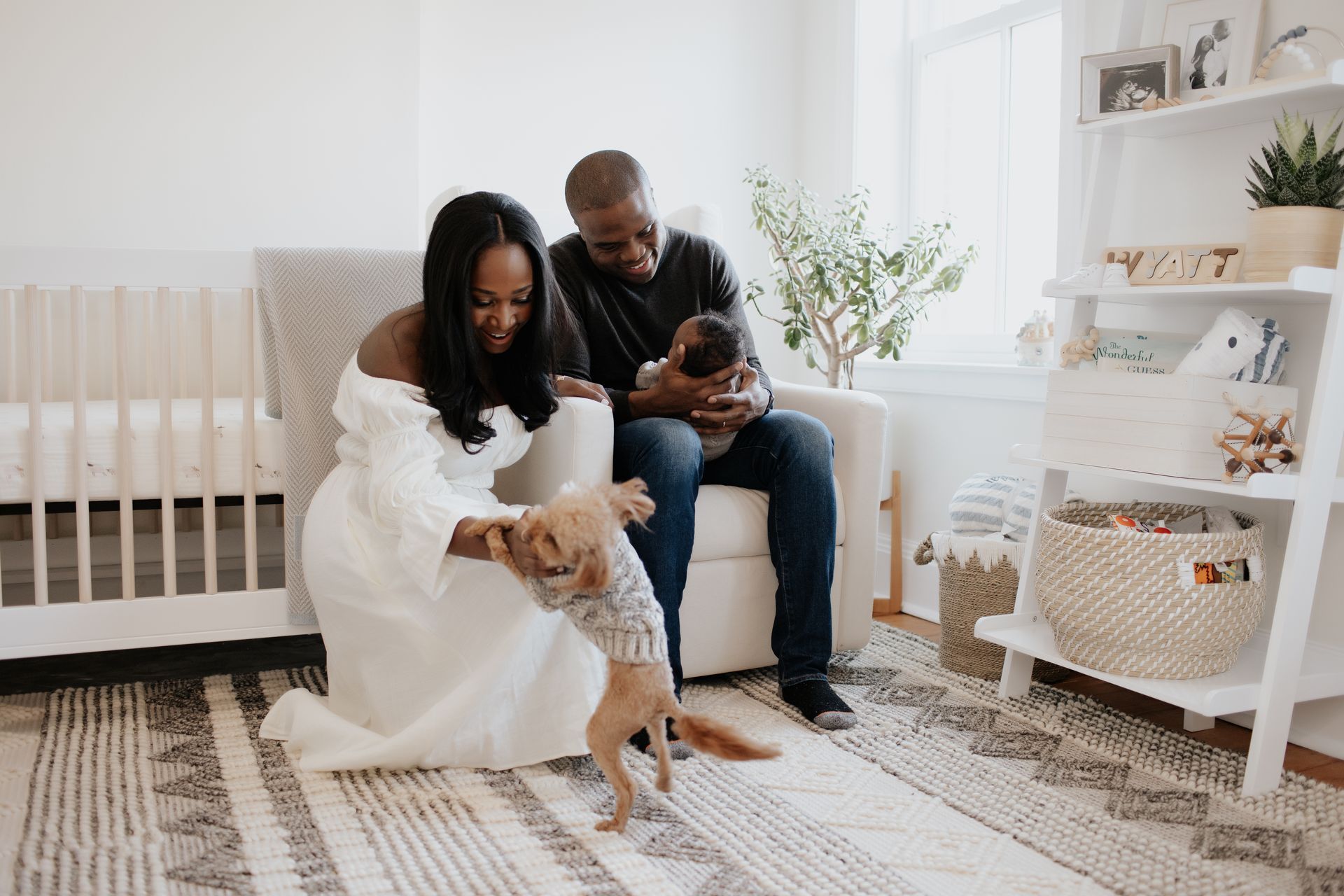 A woman is sitting in a chair holding a baby and a dog is standing next to her.