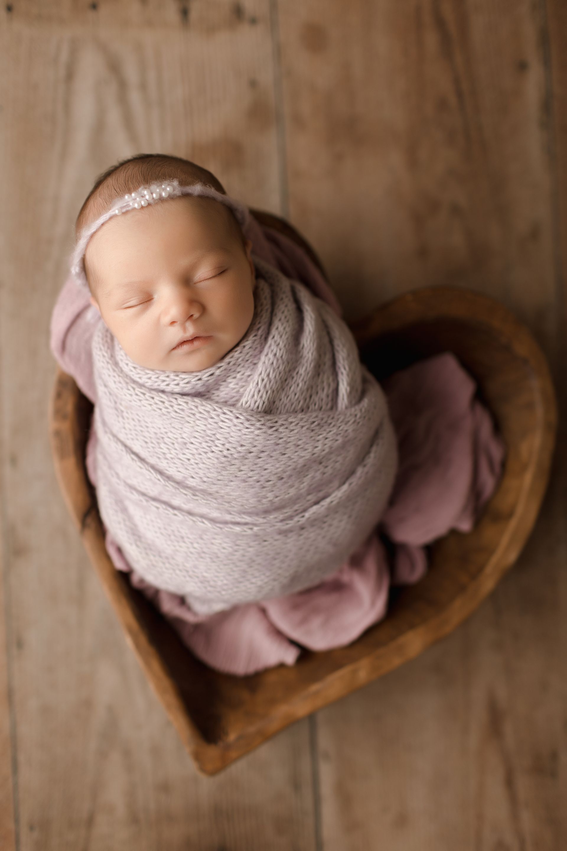 Newborn baby swaddled in lavender blanket, sleeping in a wooden heart-shaped bowl.