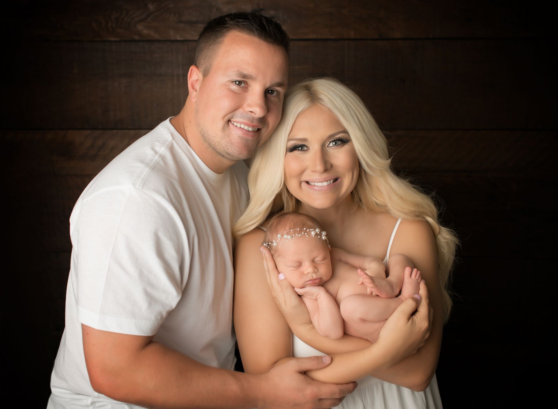 Parents holding newborn twins, smiling in front of a dark wood backdrop.
