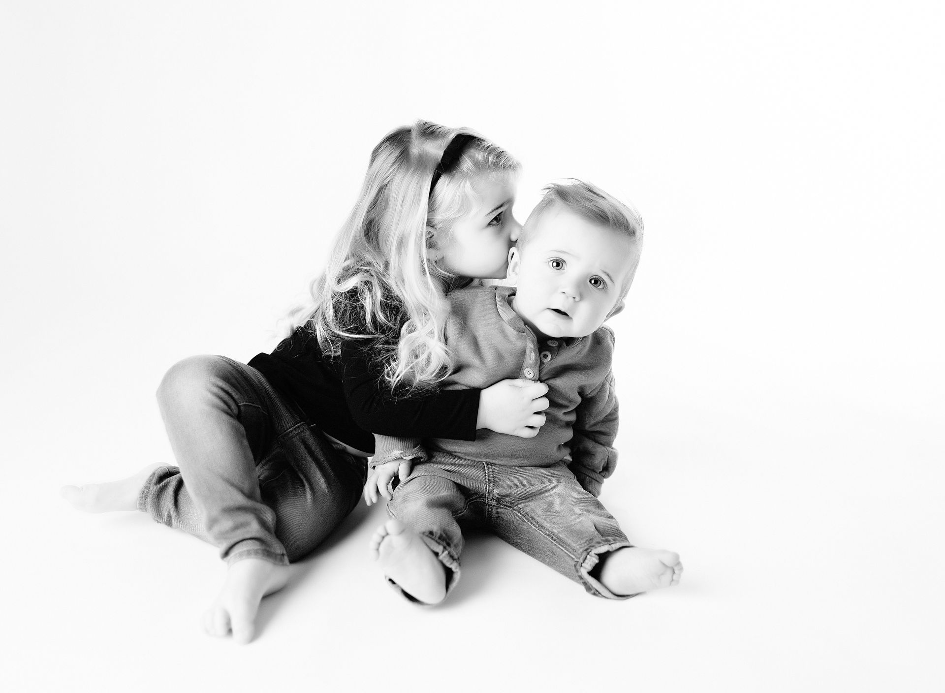 Girl kissing a baby's cheek. Both are in casual clothing, against a white backdrop.