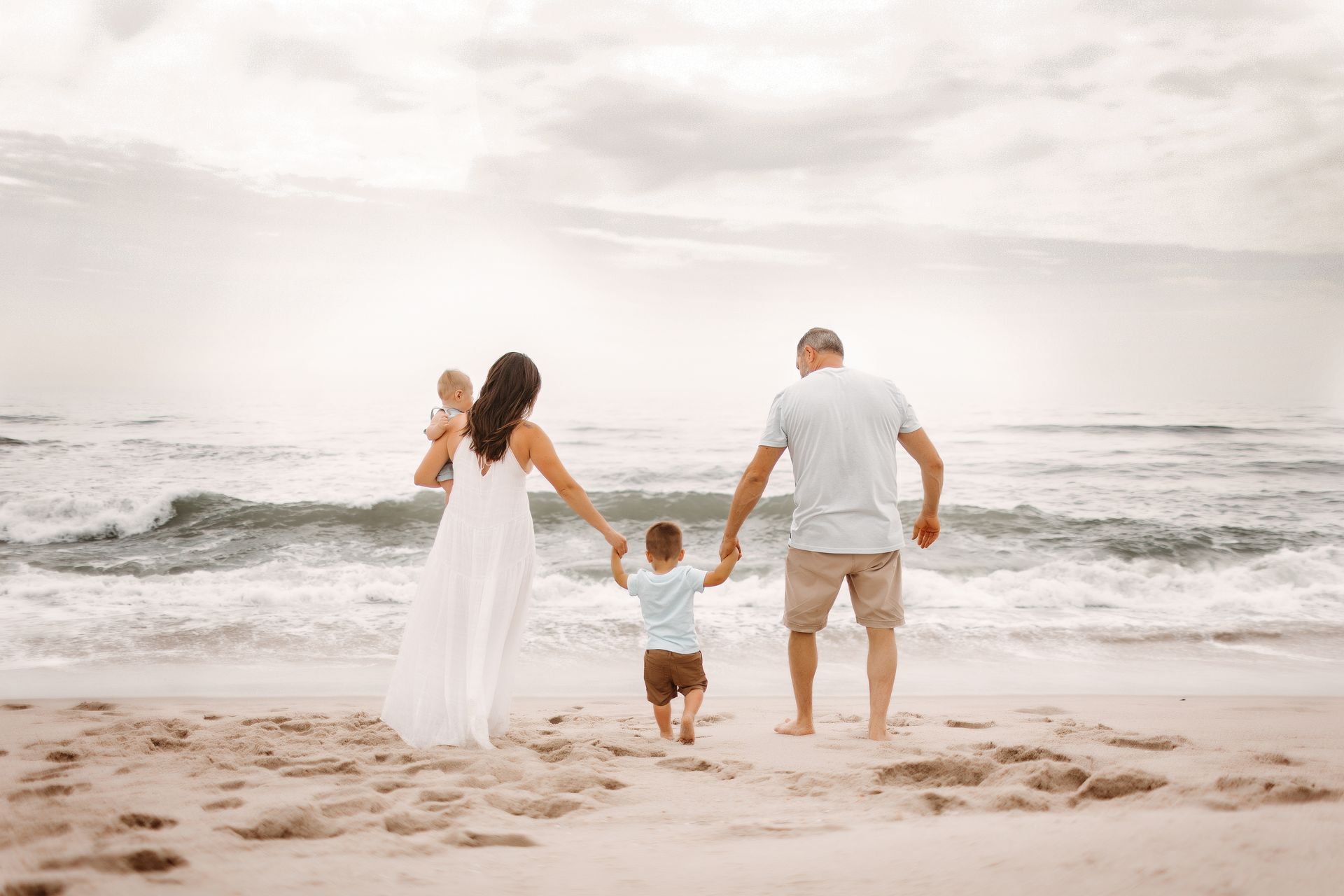 Family of four holding hands, walking on beach toward ocean, cloudy sky.