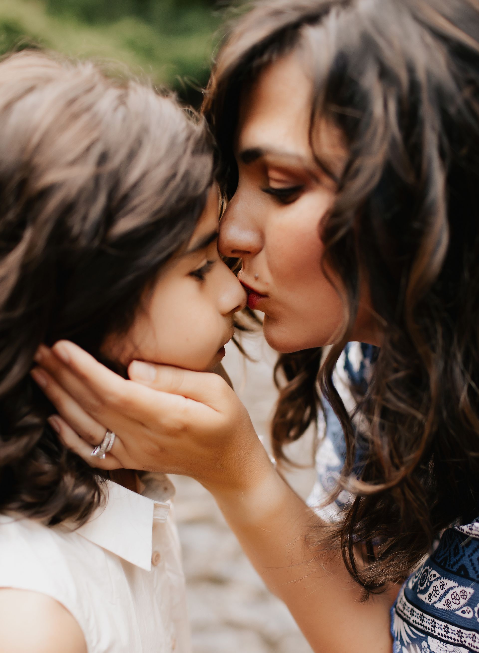 Woman kissing child's forehead; holding their face. Both have brown hair, outdoors.