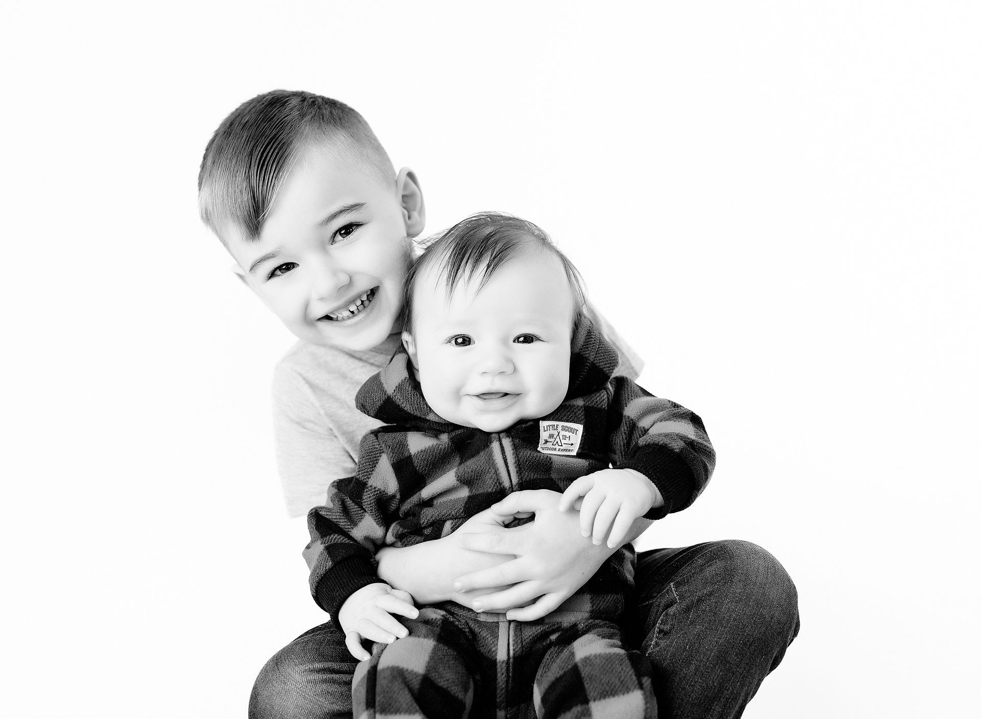 Older boy smiles, holding and looking at a baby in a plaid onesie.
