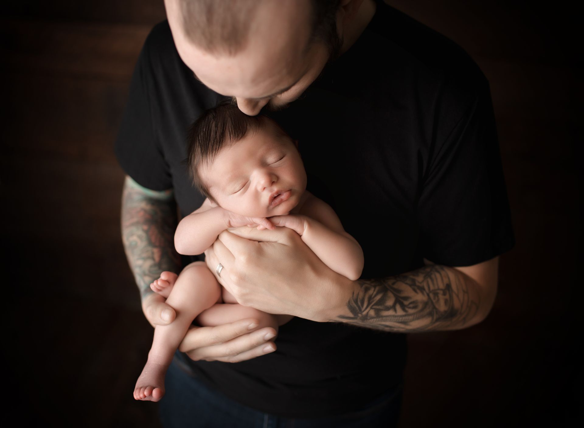 Man in black shirt cradles sleeping newborn baby. Man kisses baby's head, brown background.
