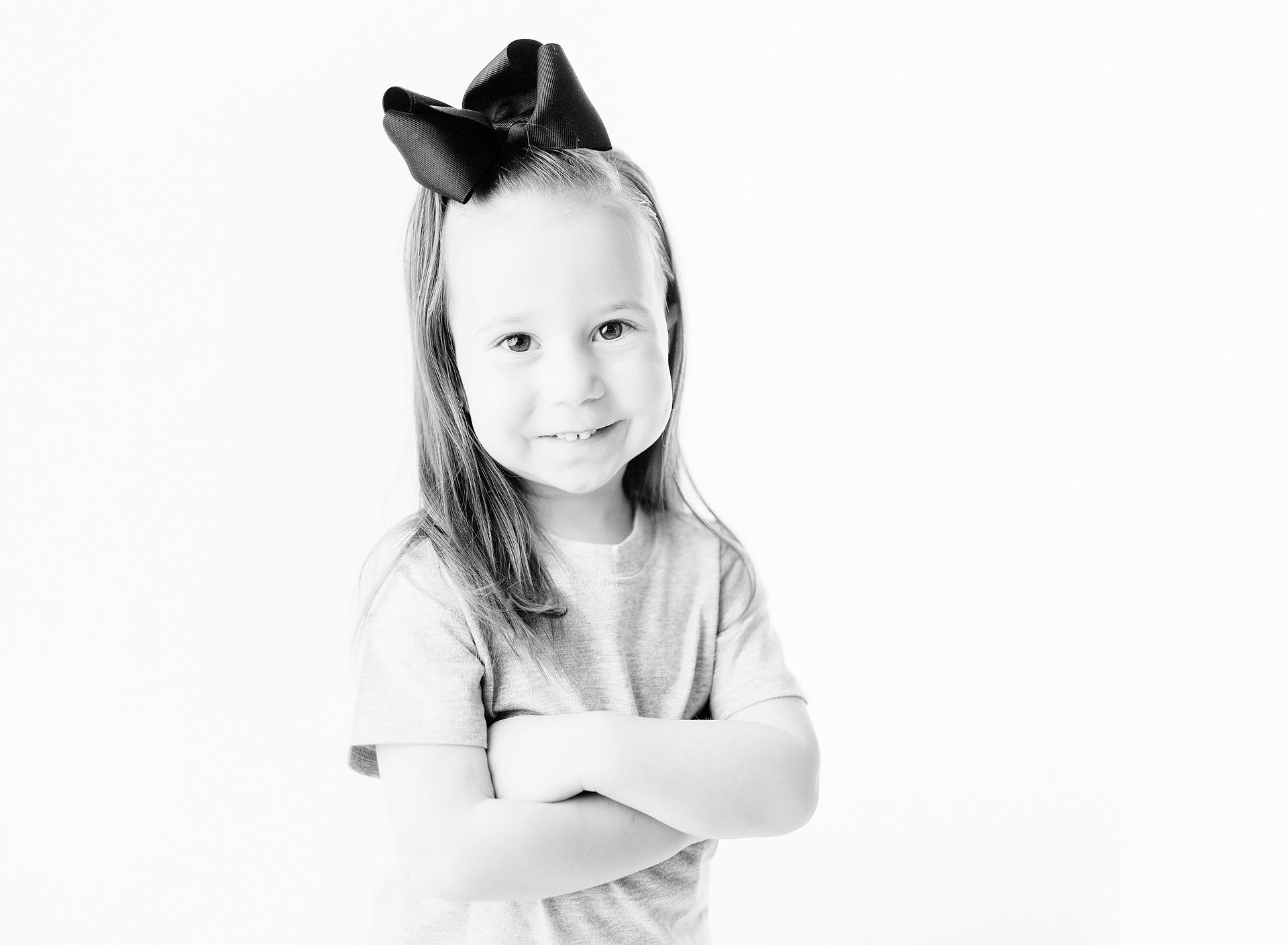 Young child with a large bow smiles, arms crossed, in a studio setting.