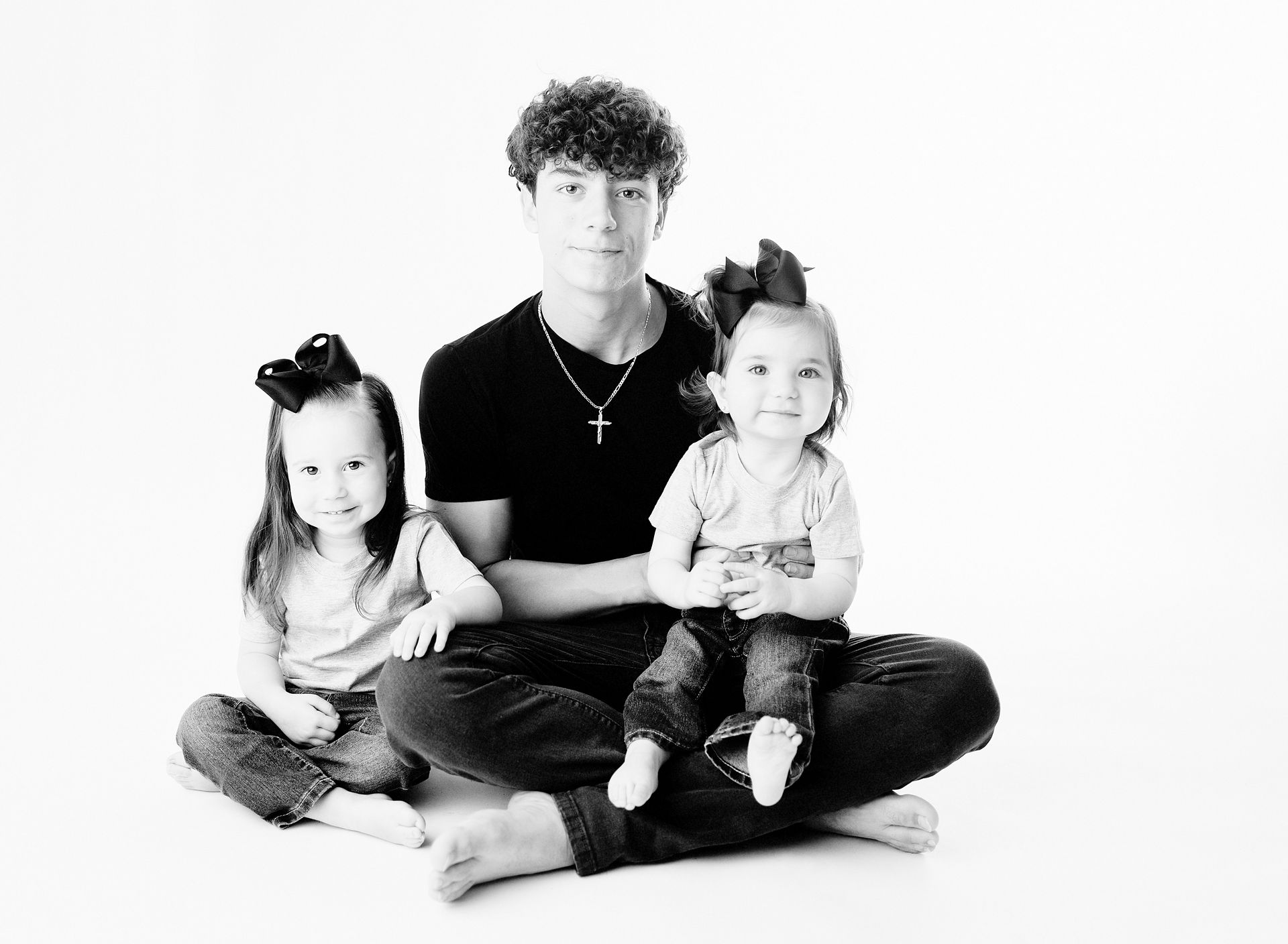 Teenage boy seated cross-legged, flanked by two young girls with bows, all in jeans, black and white studio portrait.