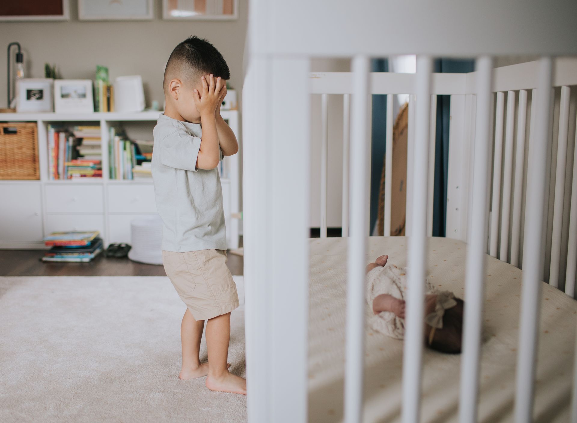 A little boy is standing in front of a crib covering his ears.