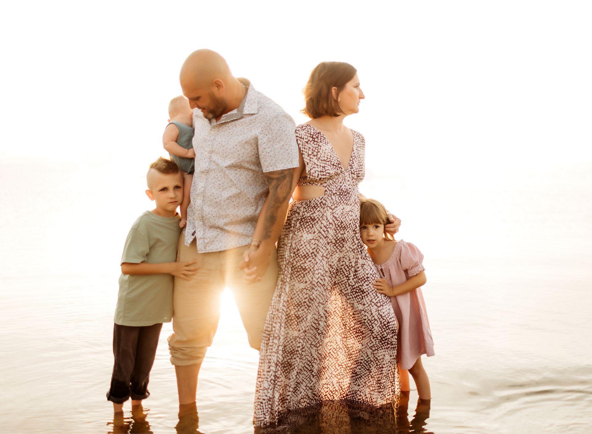 Family standing in shallow water, backlit by the sun. Parents hold hands, surrounded by three children.
