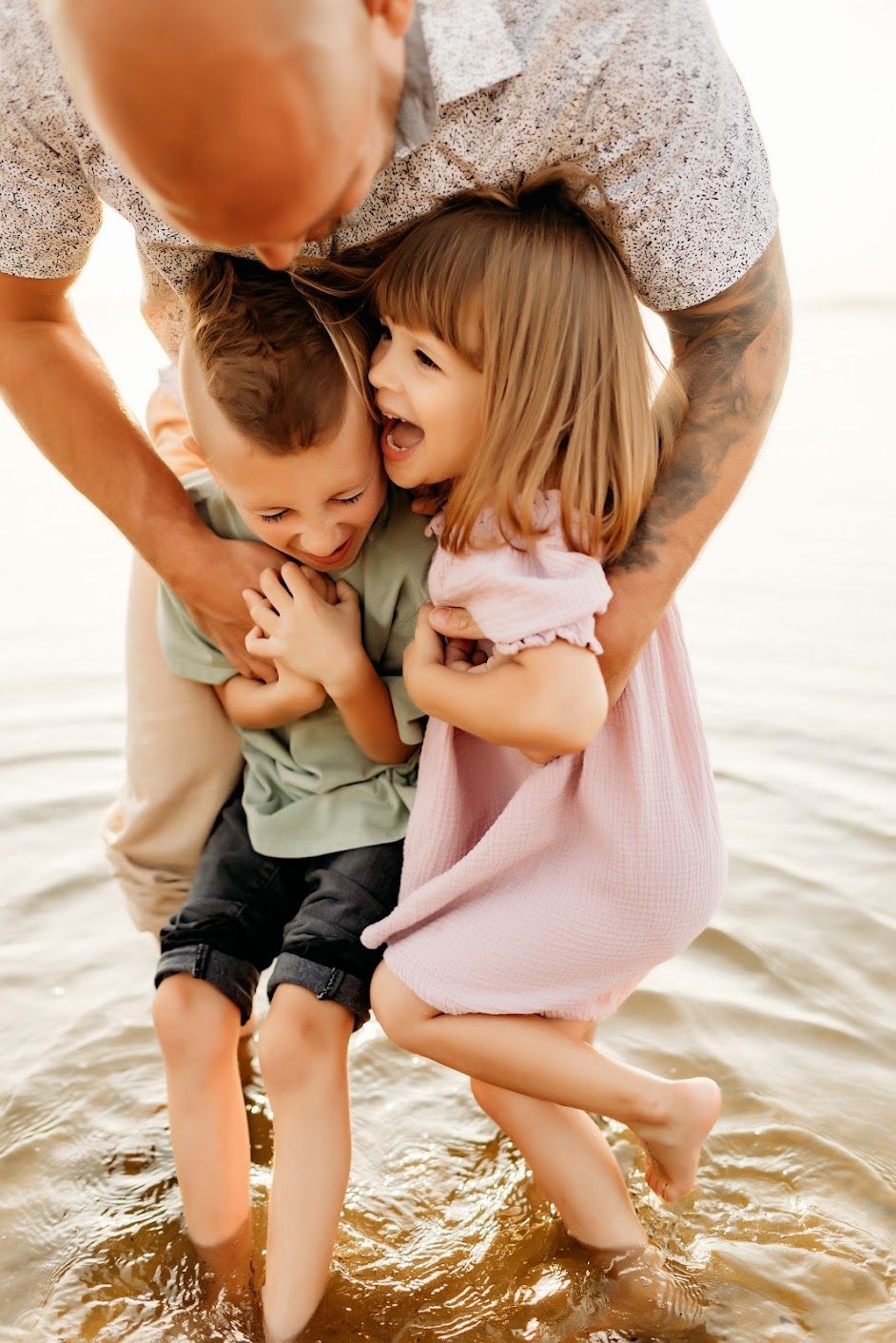 Man hugging two children in shallow water; one child laughs.