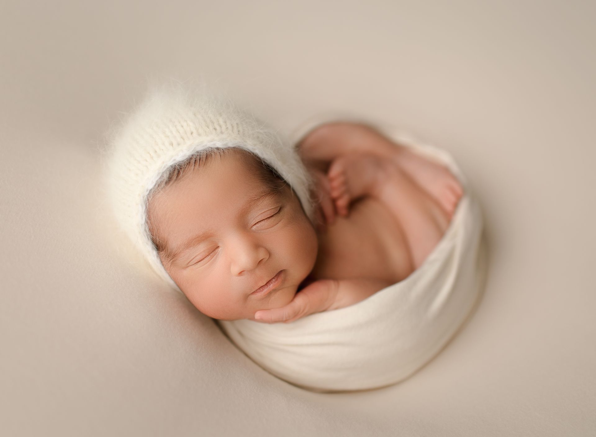 Newborn baby in a white wrap, wearing a fuzzy hat, curled up sleeping.