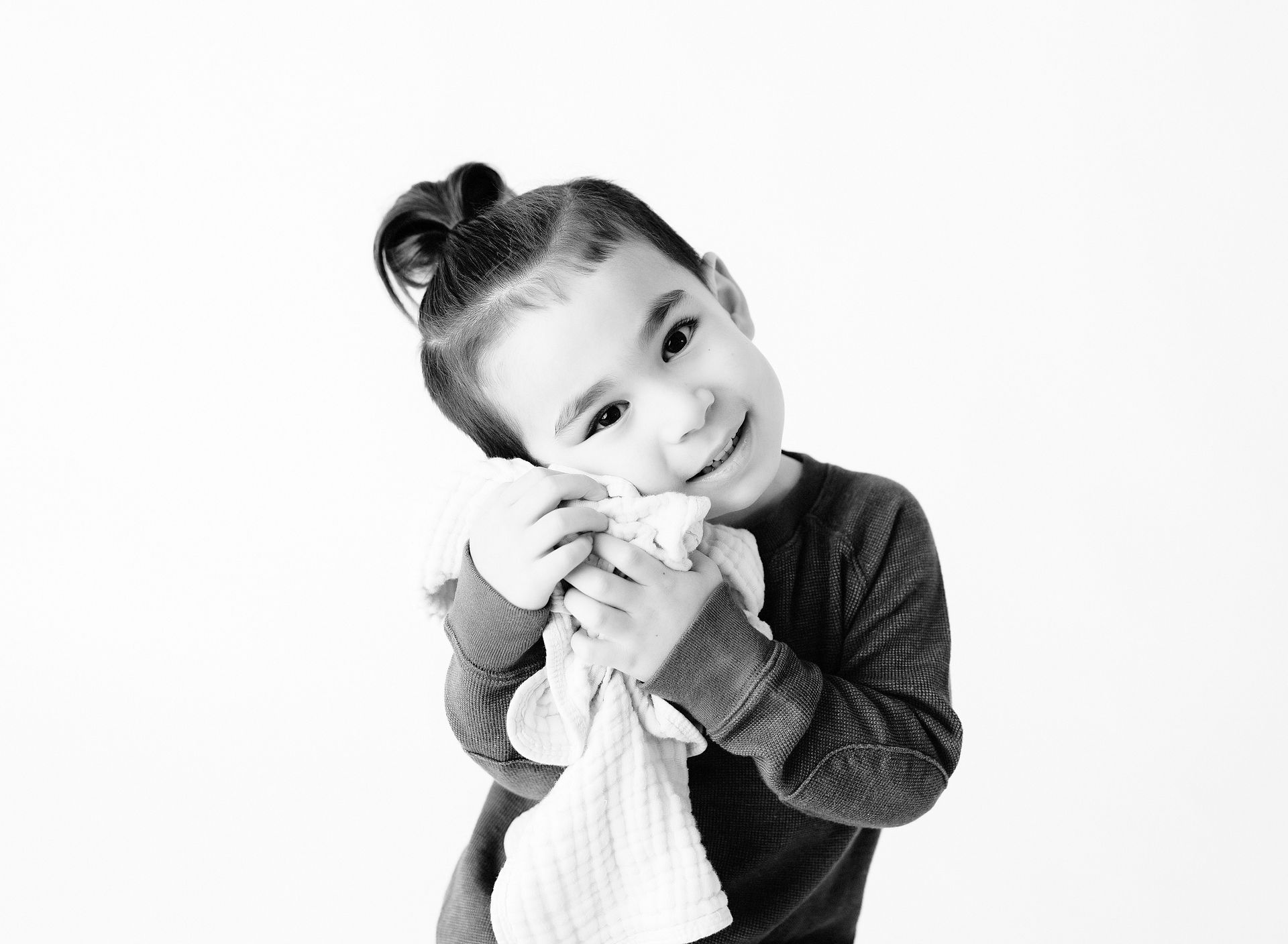 Child with bun hairstyle smiles, holding a white cloth, against a white background.