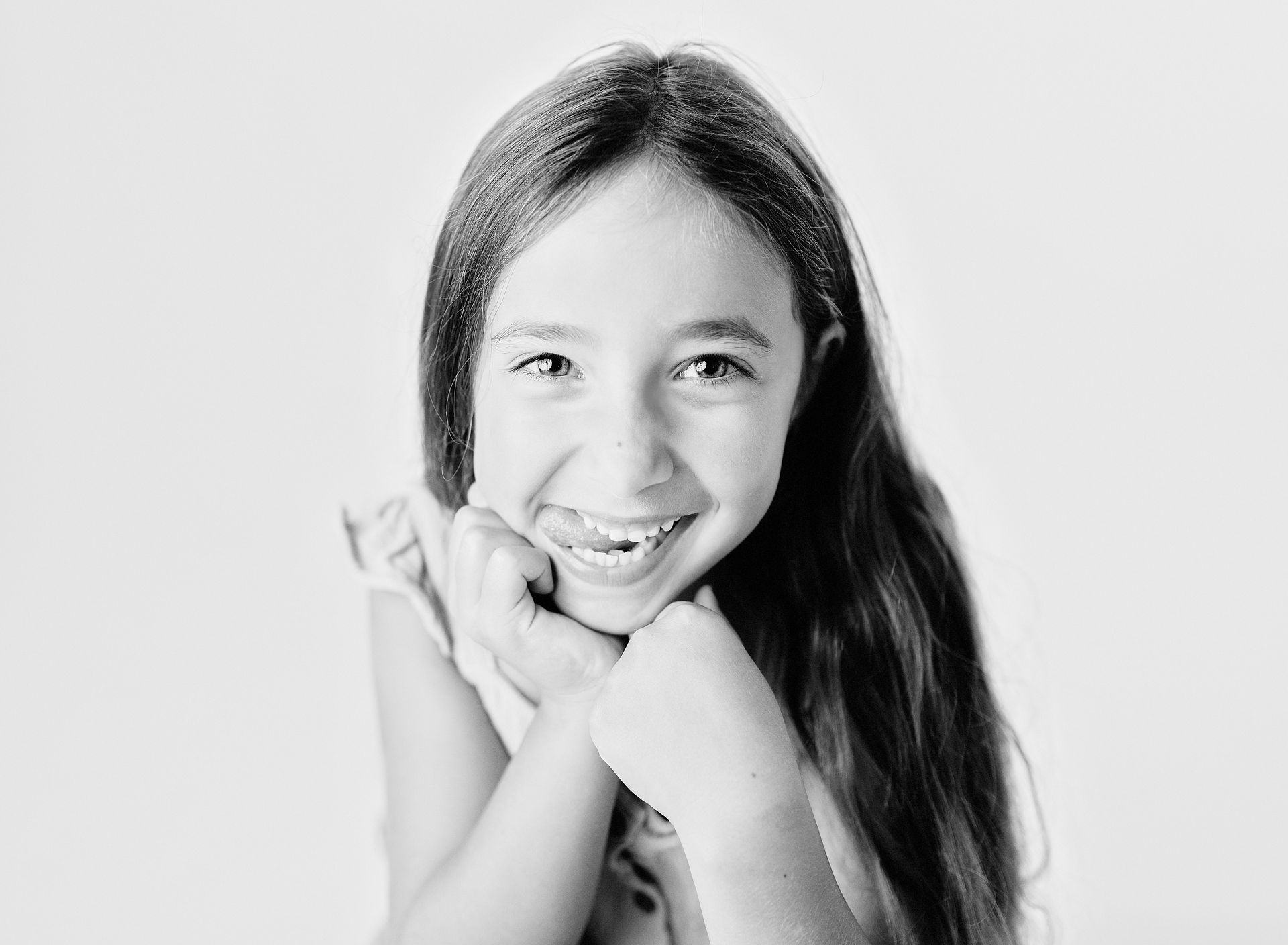Girl with long hair smiling, resting chin on clasped hands.