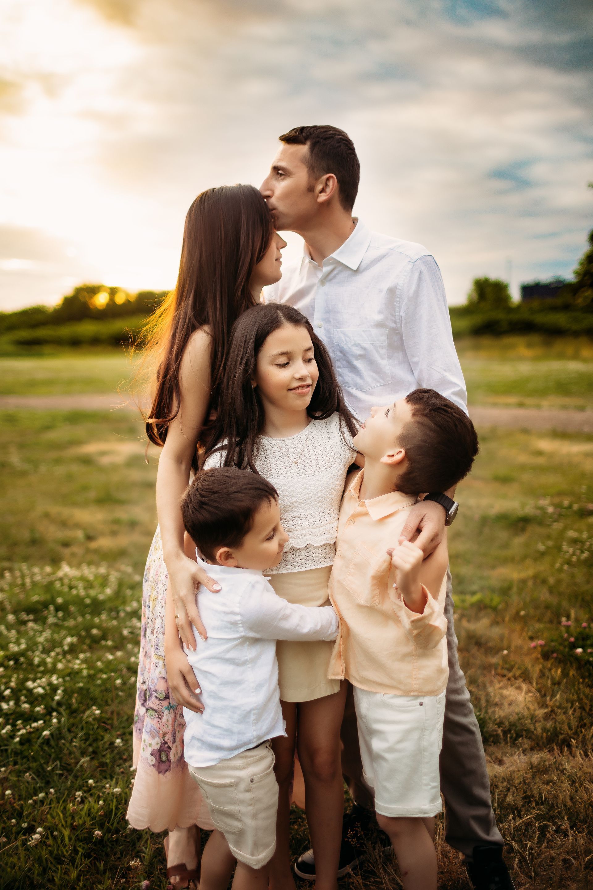 Family of five embraces in a field, man kisses woman's forehead, children smile, sunny background.